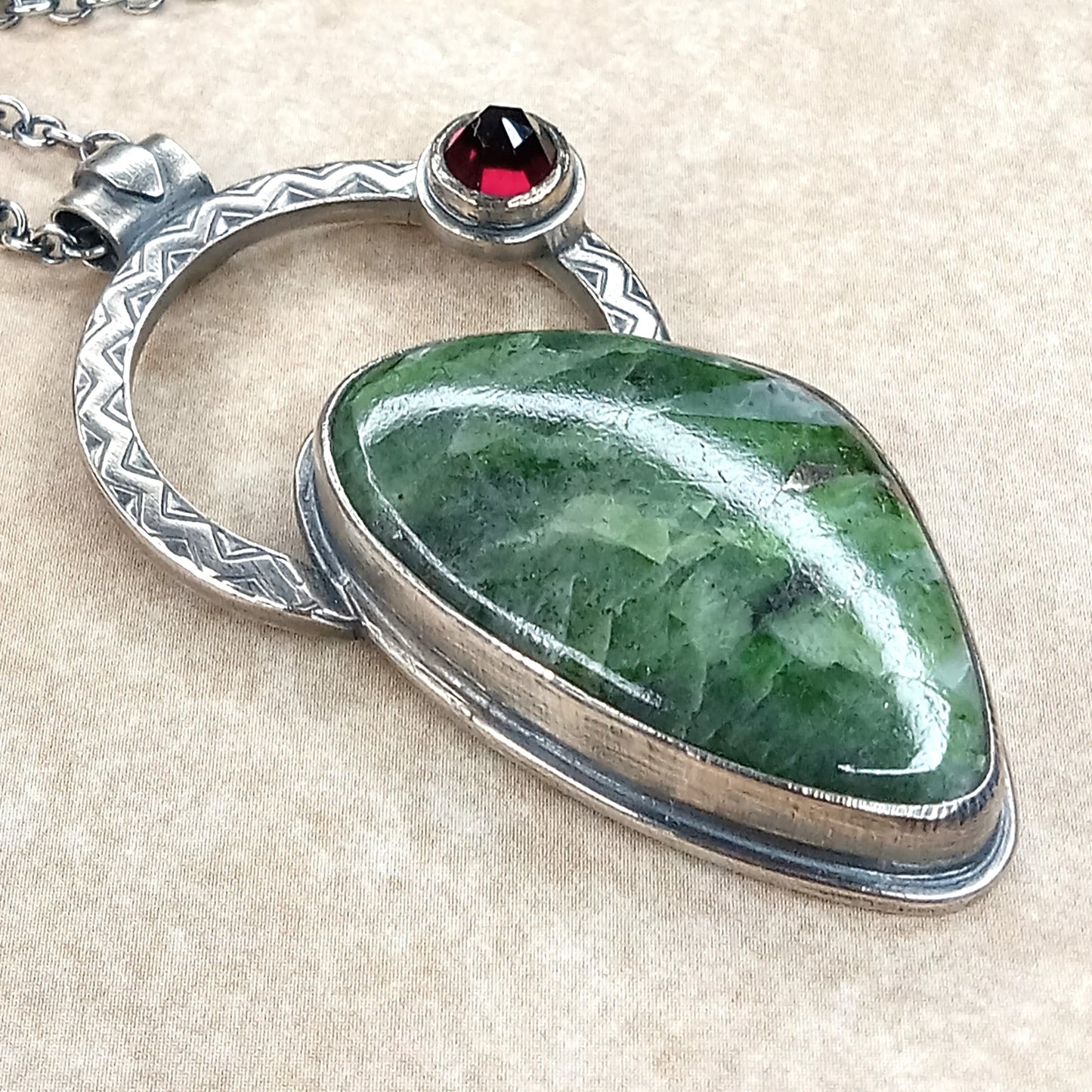 Green stone pendant with silver and red gemstone on a beige fabric background