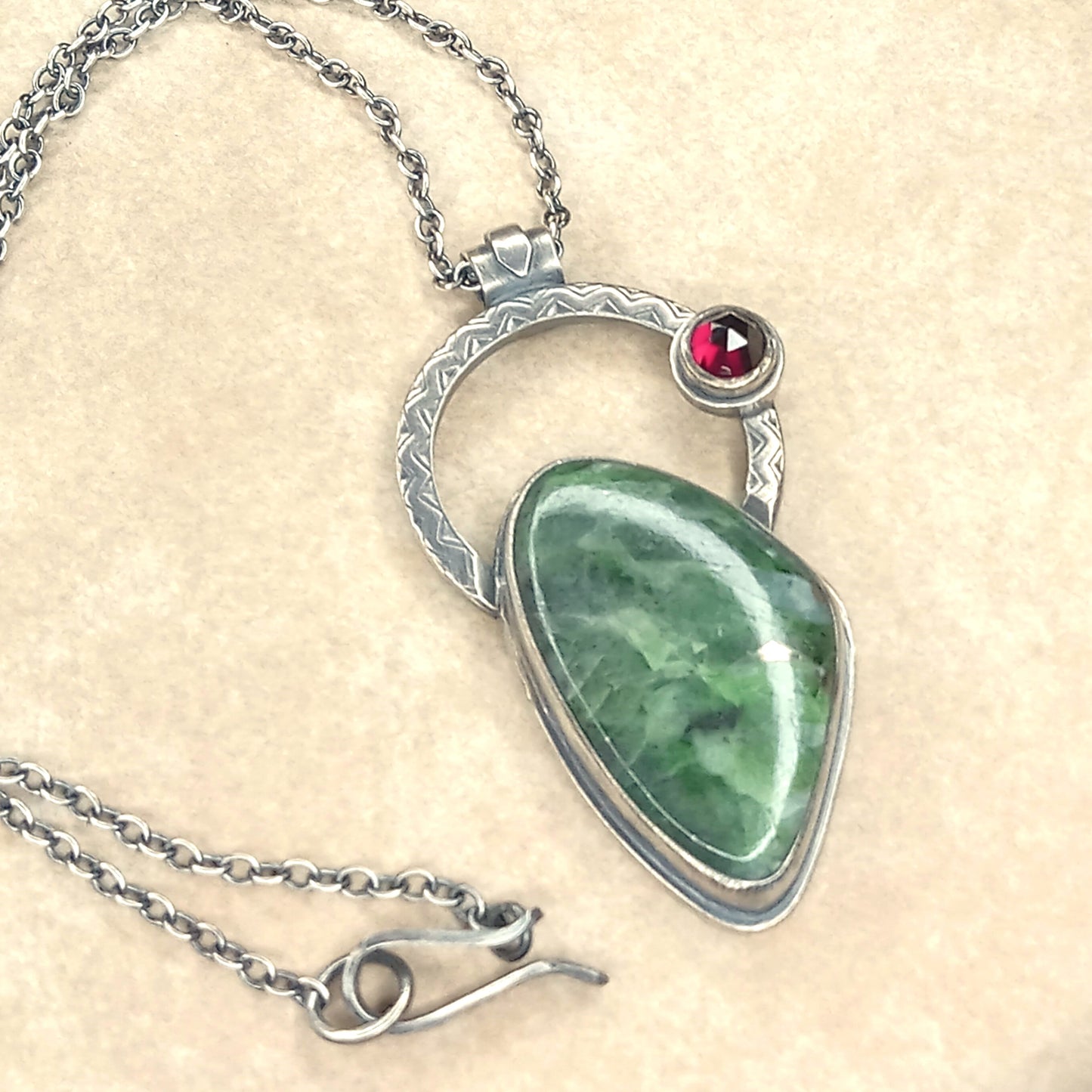 Chrome diopside and silver pendant with red garnet gemstone. Photographed on neutral sand colored background, showing clasp closure detail.