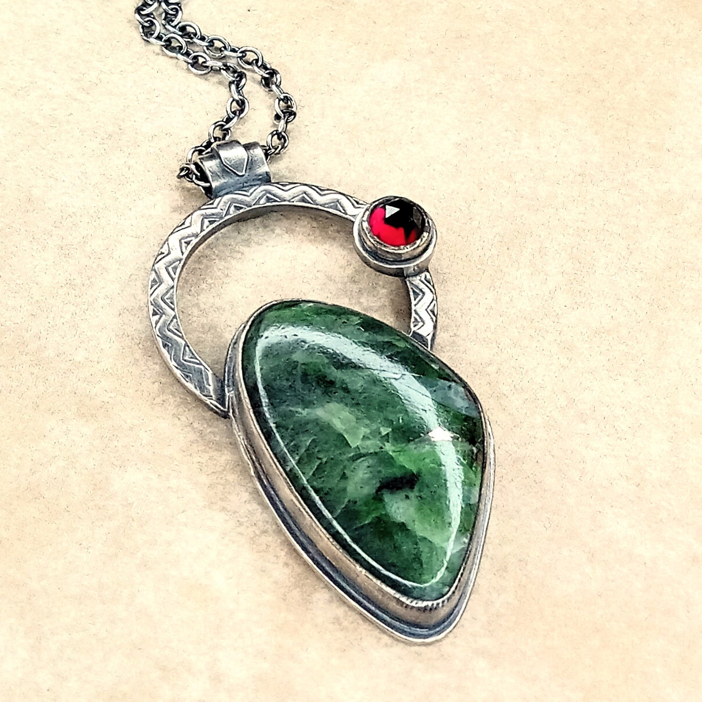 Sterling silver pendant featuring a green chrome diopside cabochon with a zigzag halo and rose cut garnet accent on 18-inch chain.