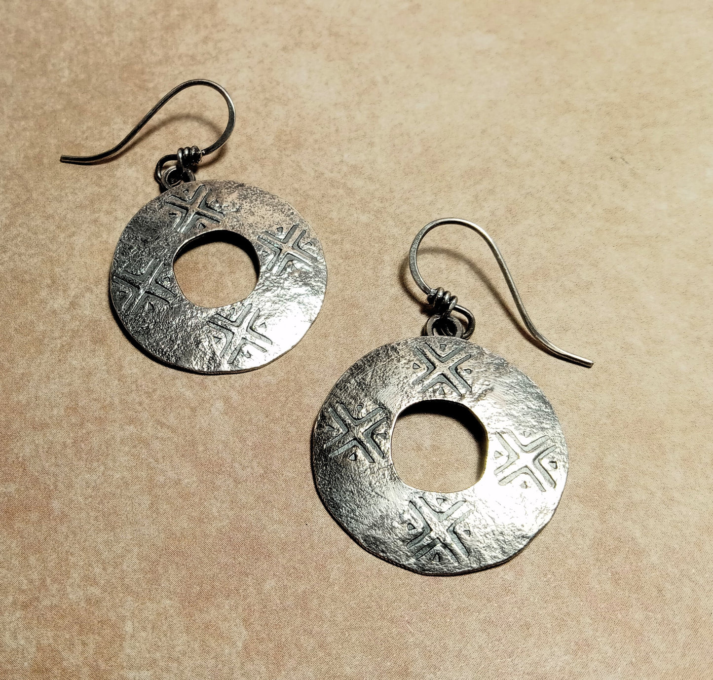 Pair of rustic argentium silver round coin style earrings by Mocahete on a beige background.