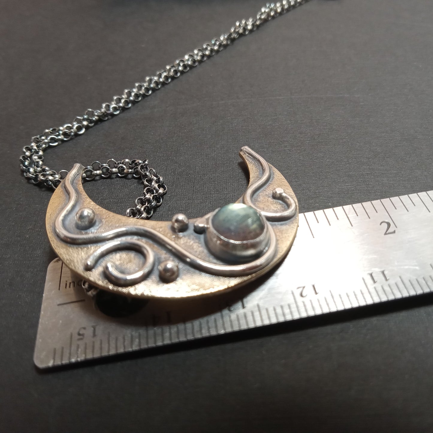 Silver, bronze and labradorite crescent moon necklace on a black surface with a ruler for scale.
