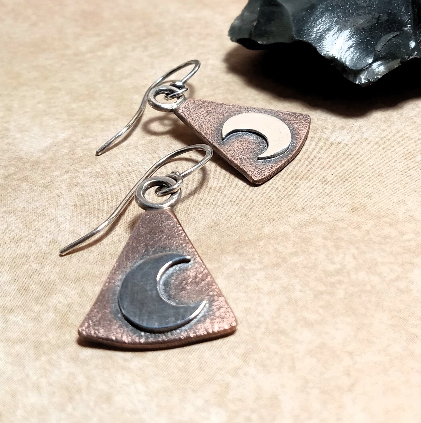 Copper triangle earrings with moon symbols on a beige surface