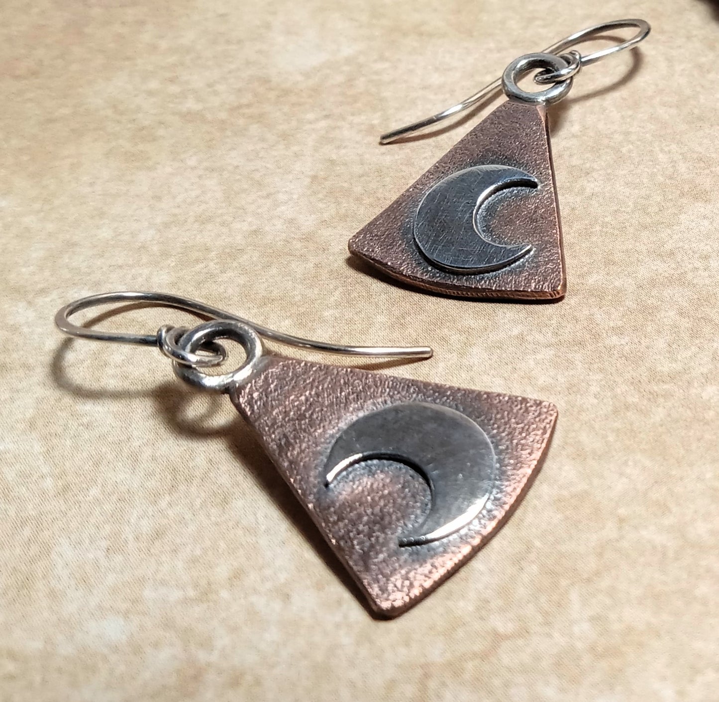 Triangular earrings in mixed metals with moon designs on a beige background.