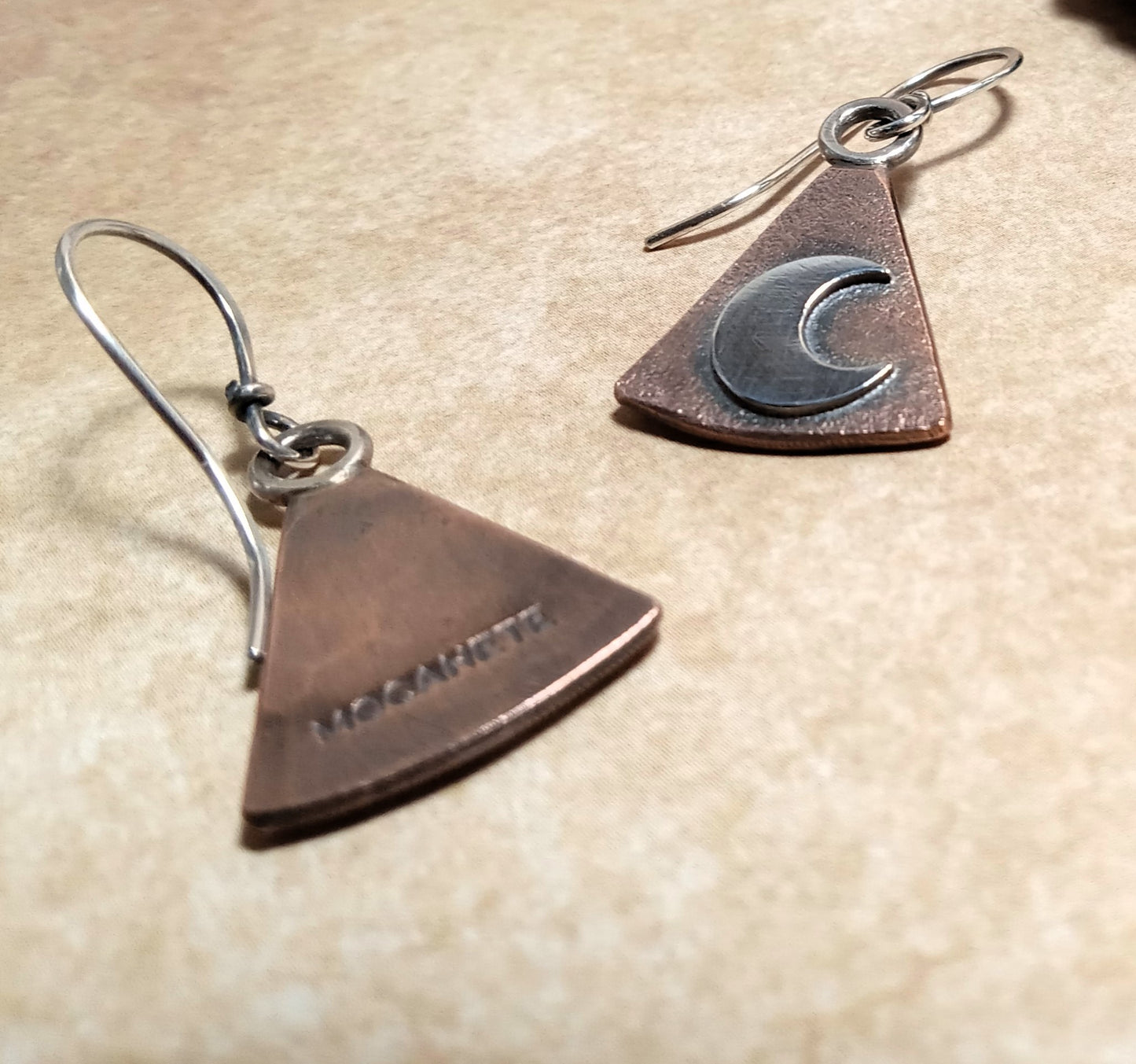 Pair of handcrafted copper and silver moon earrings showing makers mark on reverse side on a beige surface.
