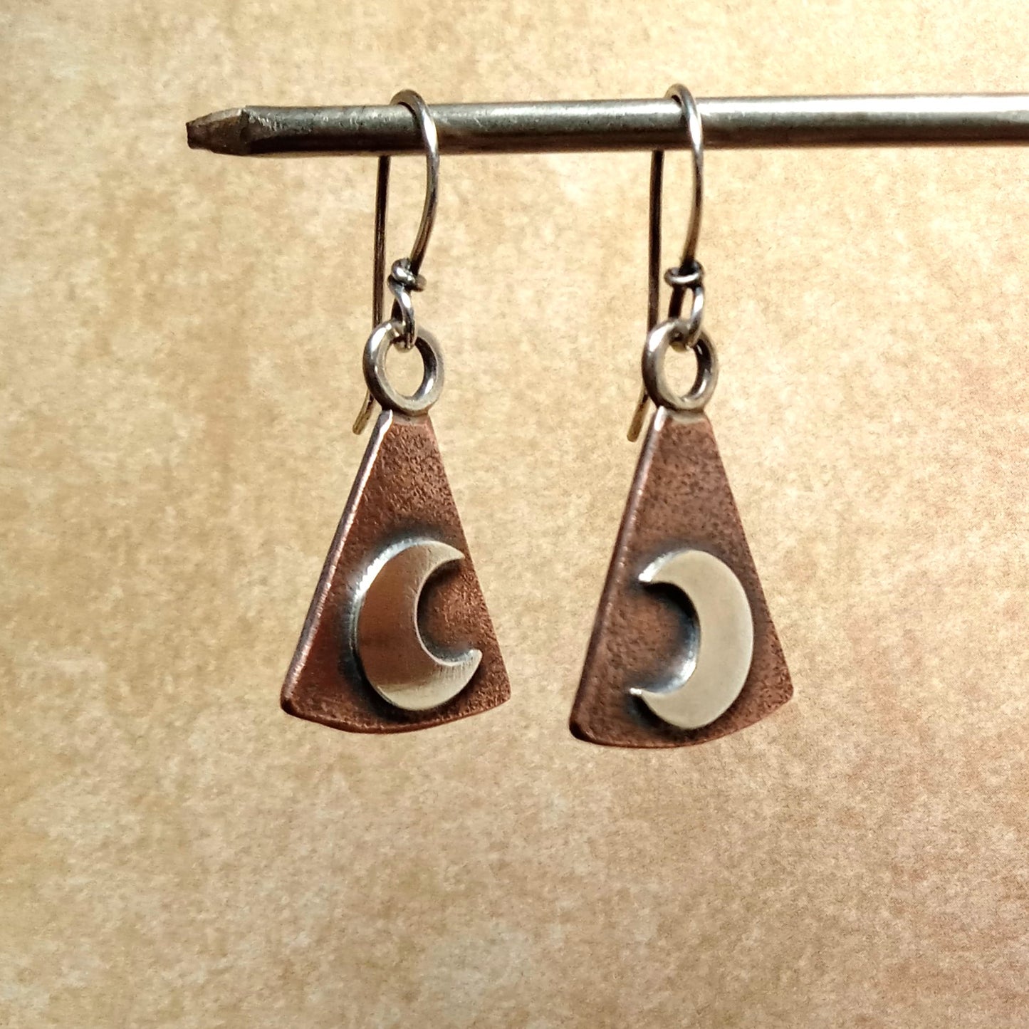 Pair of earrings with triangular copper and silver crescent moon design on a beige background.