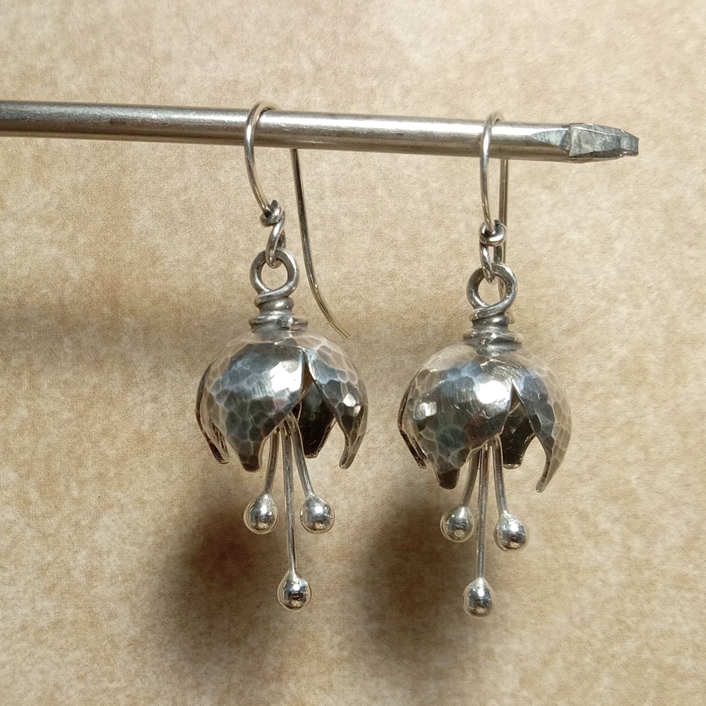 Handcrafted petite bell flower earrings in Argentium sterling silver with hammered texture and three balled silver stamens that create a soft tinkling sound. Lightweight, oxidized, and polished for depth and shine. Suspended on stand.