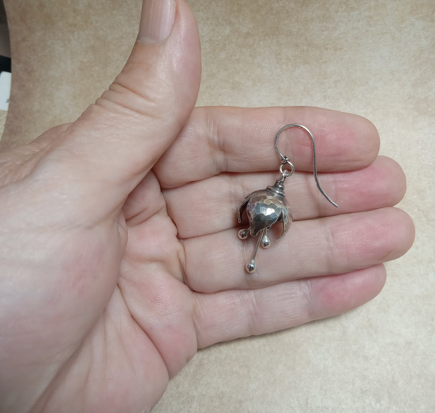 Silver bell flower earrings held in a hand against a beige background