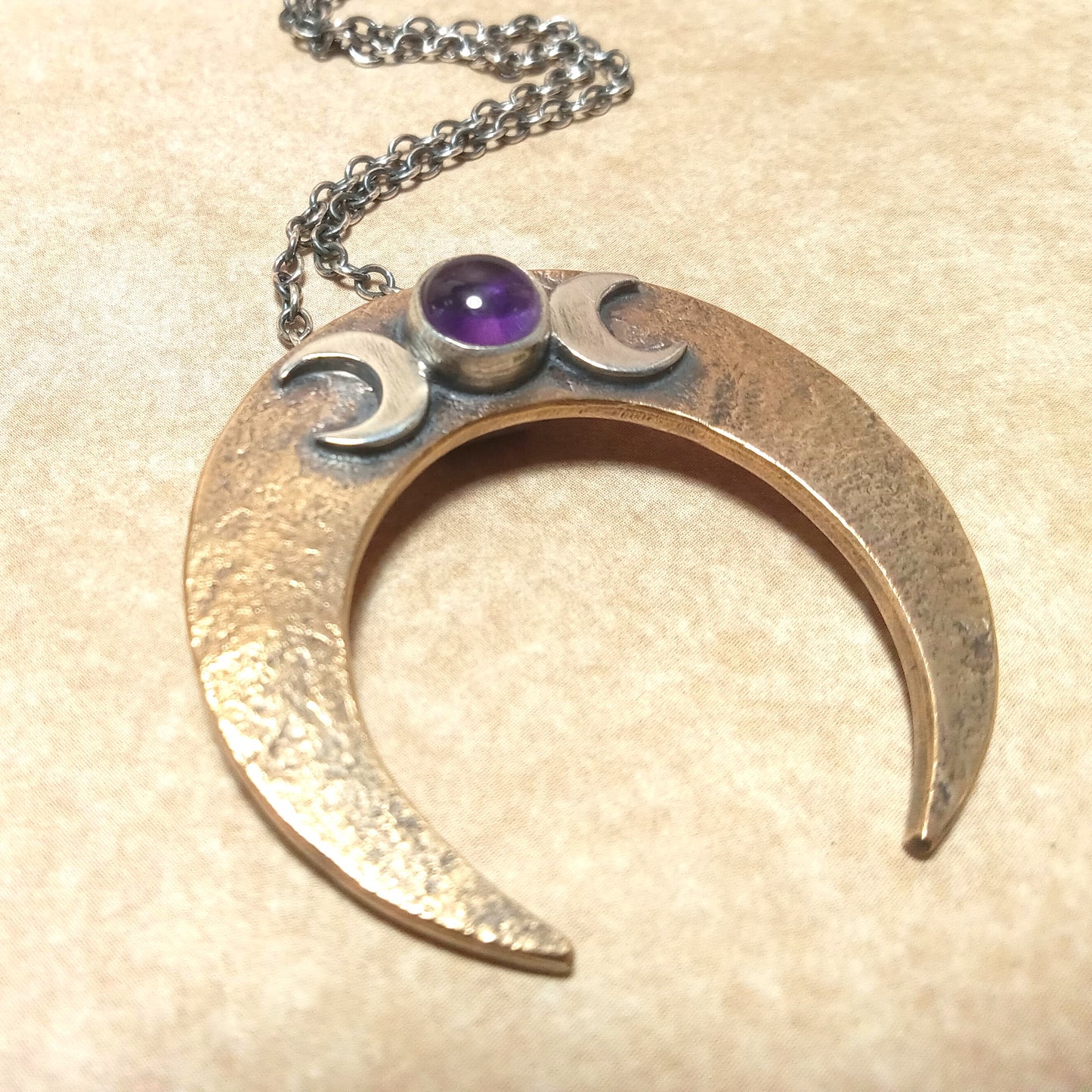 Crescent moon-shaped pendant with two small moons and an amethyst gemstone on a beige background.