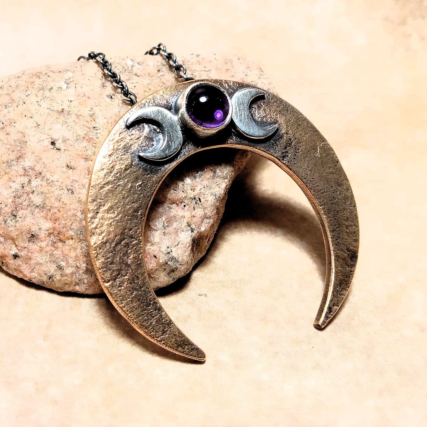 Downward facing crescent moon-shaped pendant in bronze with sterling silver and a purple amethyst on a beige stone background.