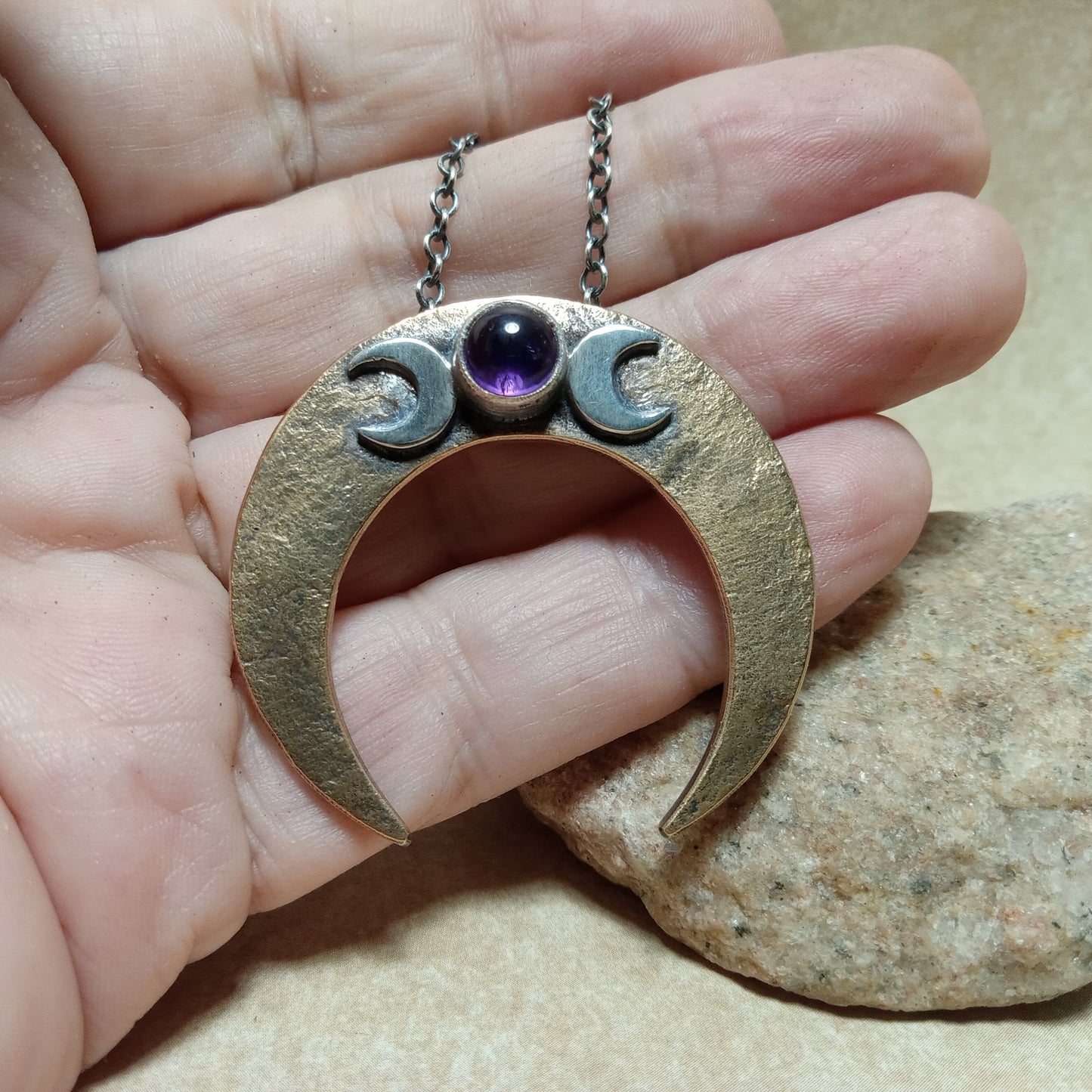 Hand holding a bronze and silver crescent moon pendant with a purple gemstone against a stone background.