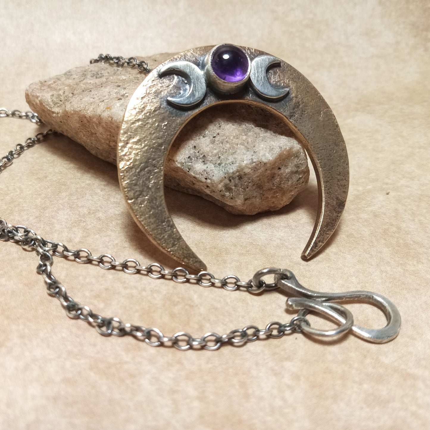 Bronze and Silver pagan crescent moon pendant with a amethyst gemstone, showing chain and handmade findings on a beige background.