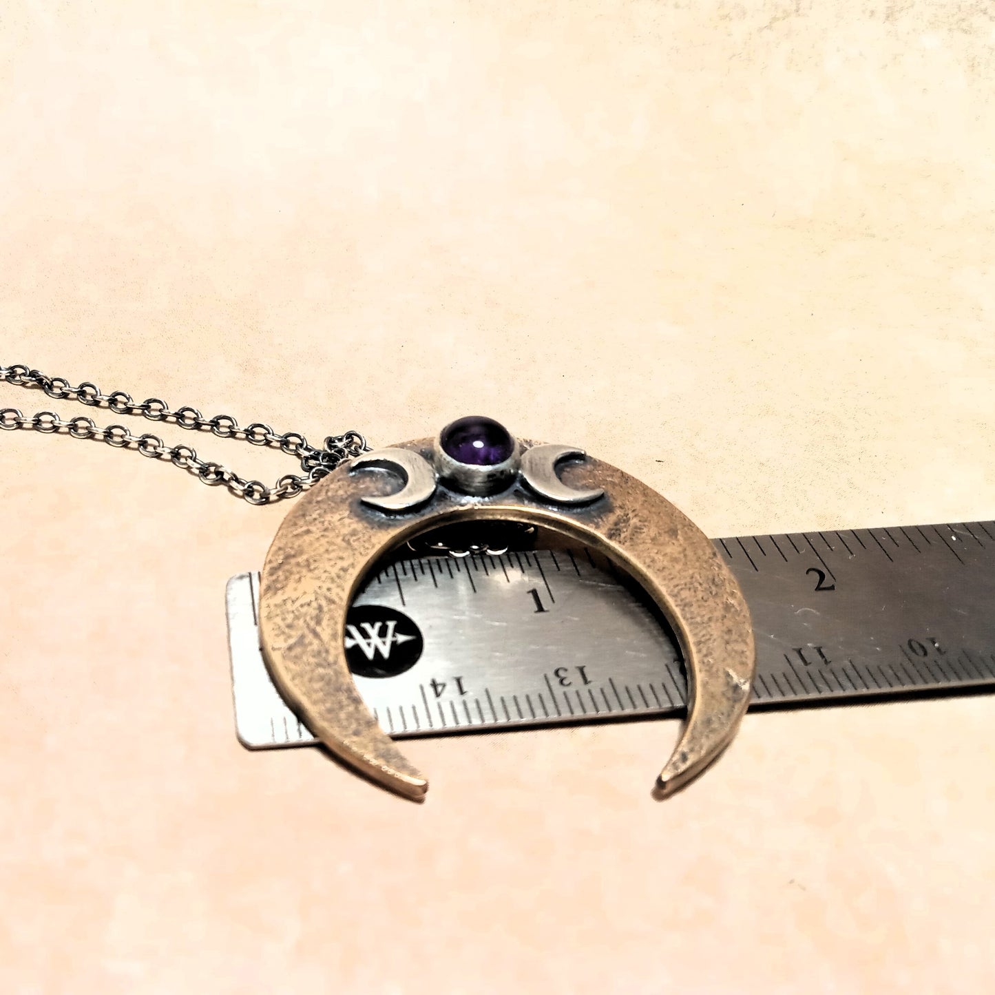 Bronze necklace with a crescent moon design and purple gemstone, displayed on a beige surface with a ruler for scale.