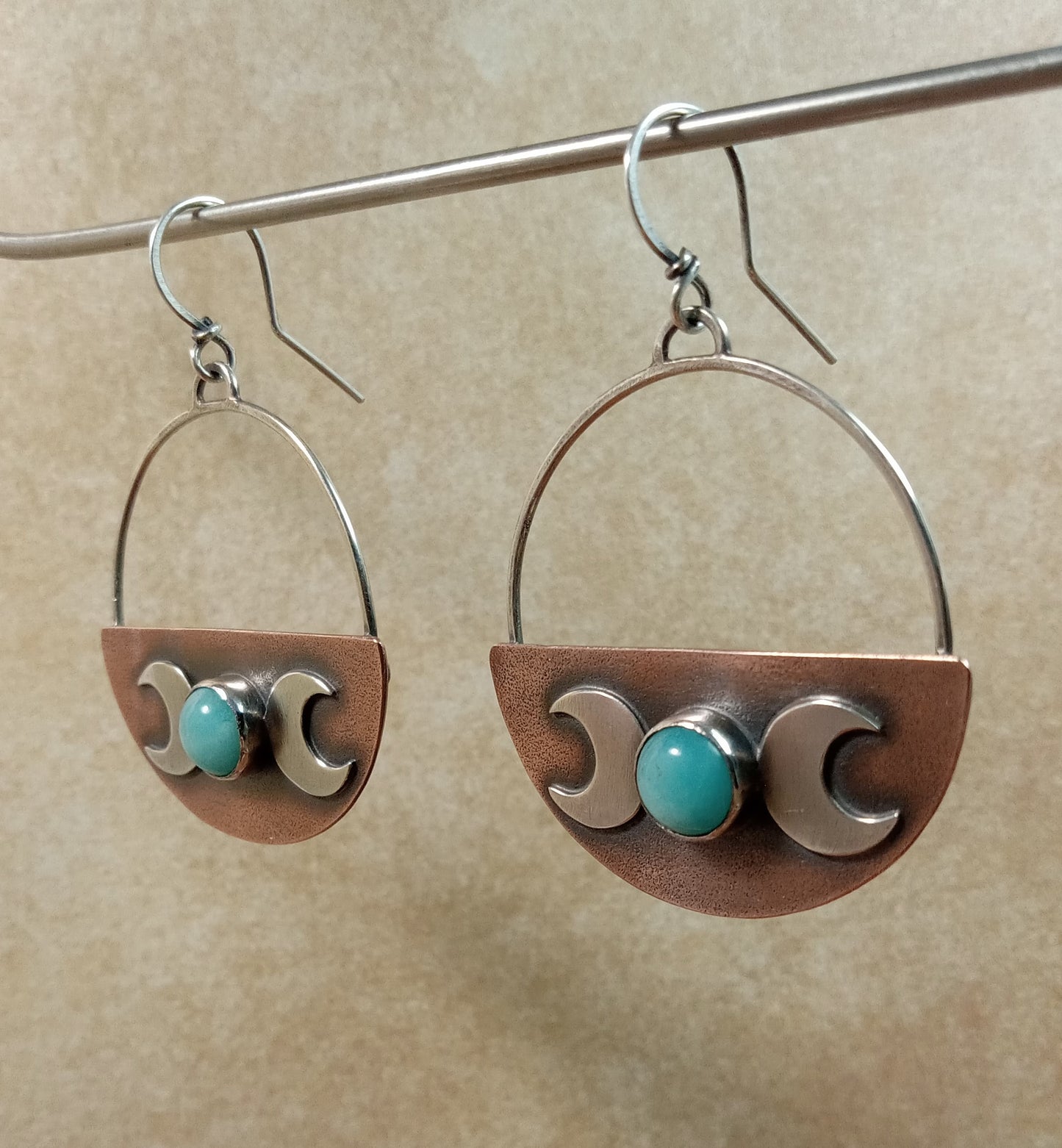 Pair of triple goddess earrings with copper and silver design, amazonite cabochons hanging from stand on a beige background.