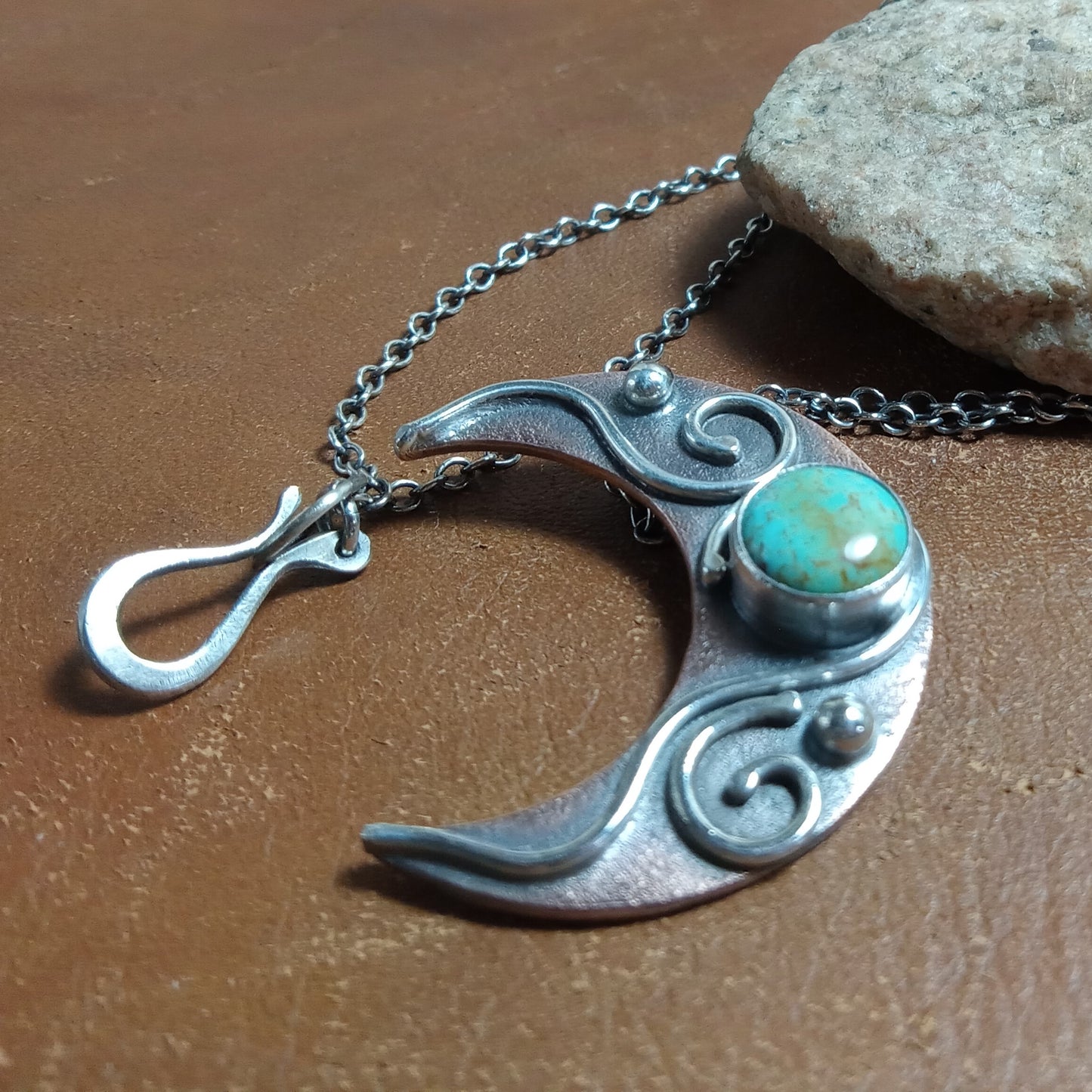 Copper and Silver crescent moon pendant with turquoise stone jewelry by Mocahete on a brown surface.