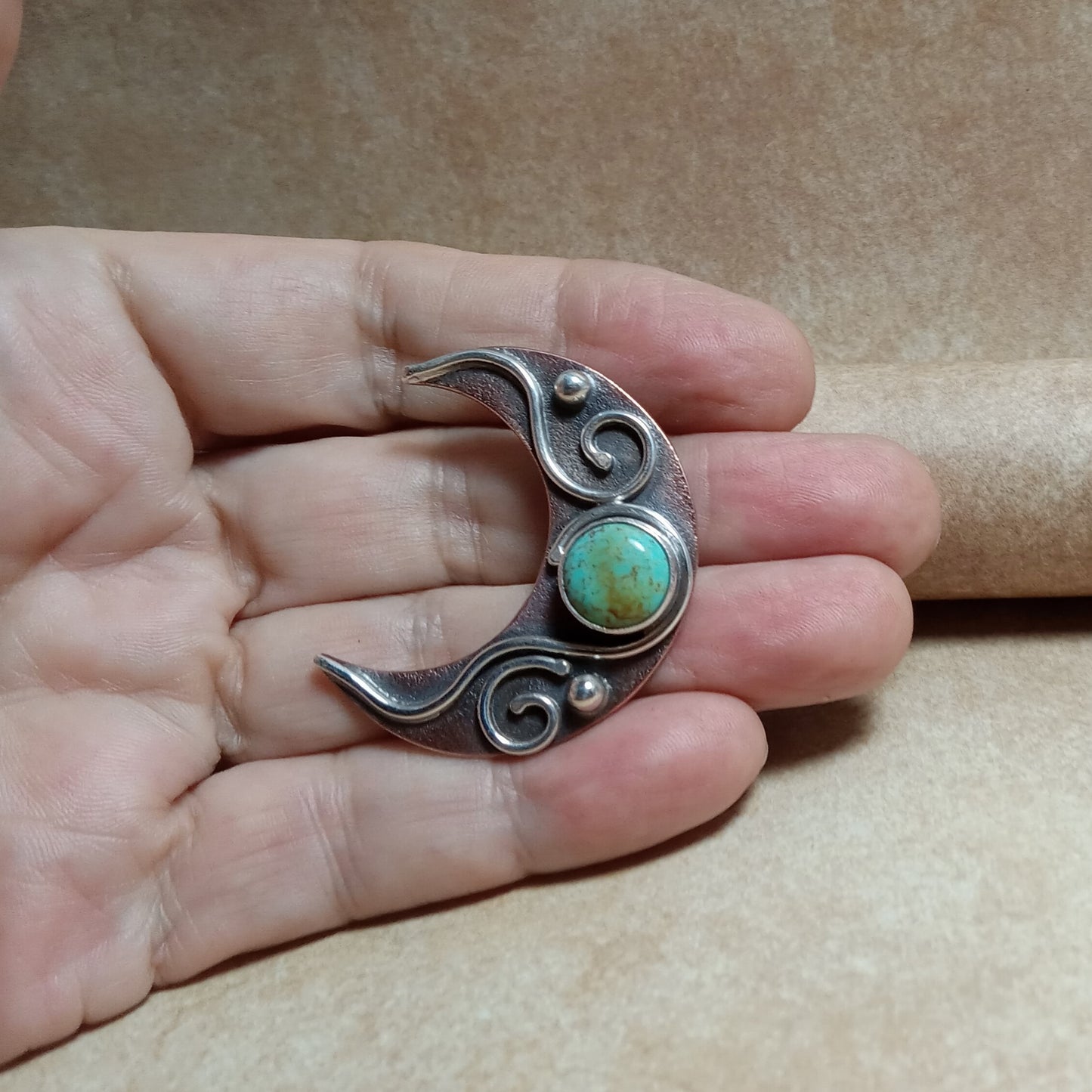 Silver and copper crescent moon pendant with turquoise stone held in a hand against a brown background, jewelry by Mocahete.