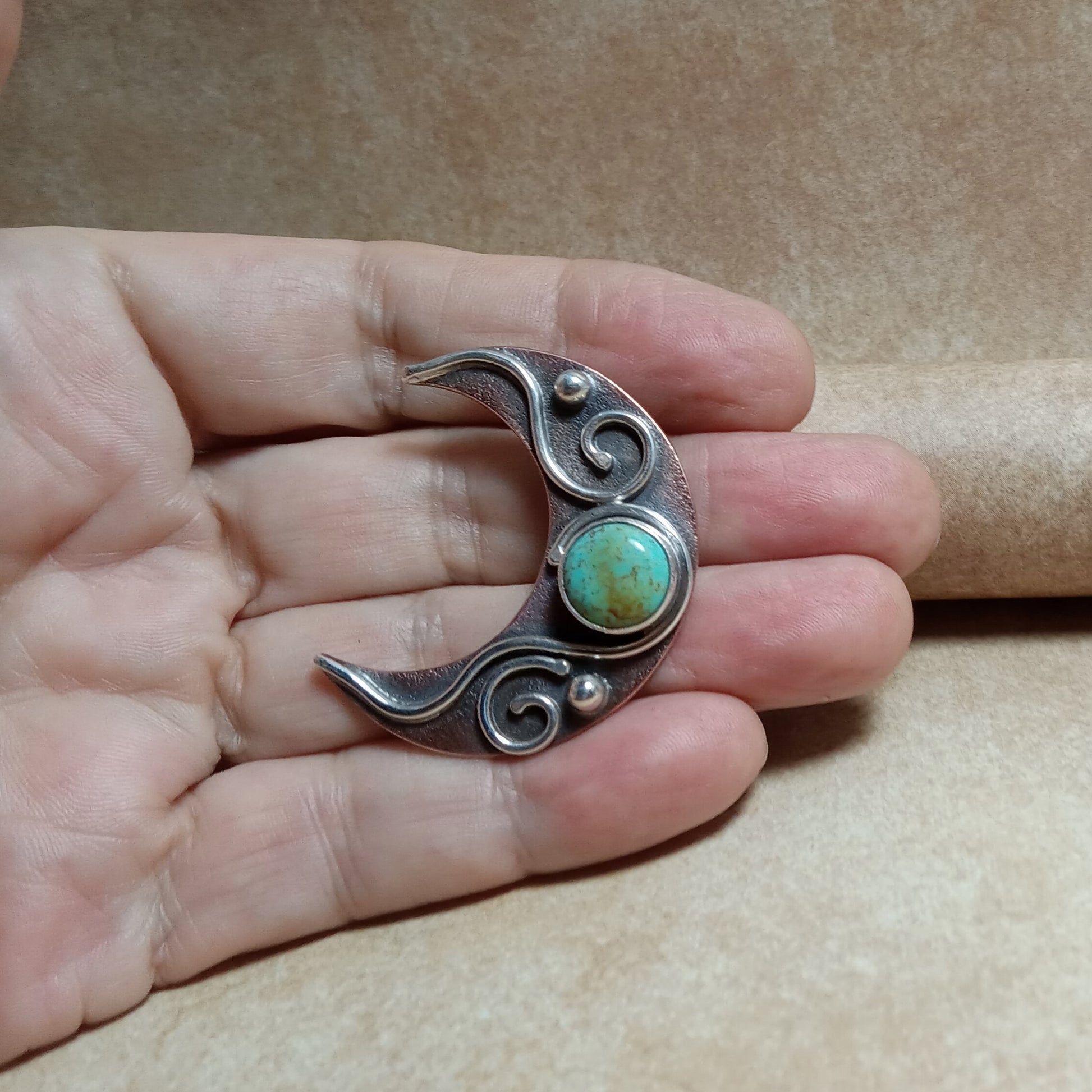 Silver and copper crescent moon pendant with turquoise stone held in a hand against a brown background, jewelry by Mocahete.