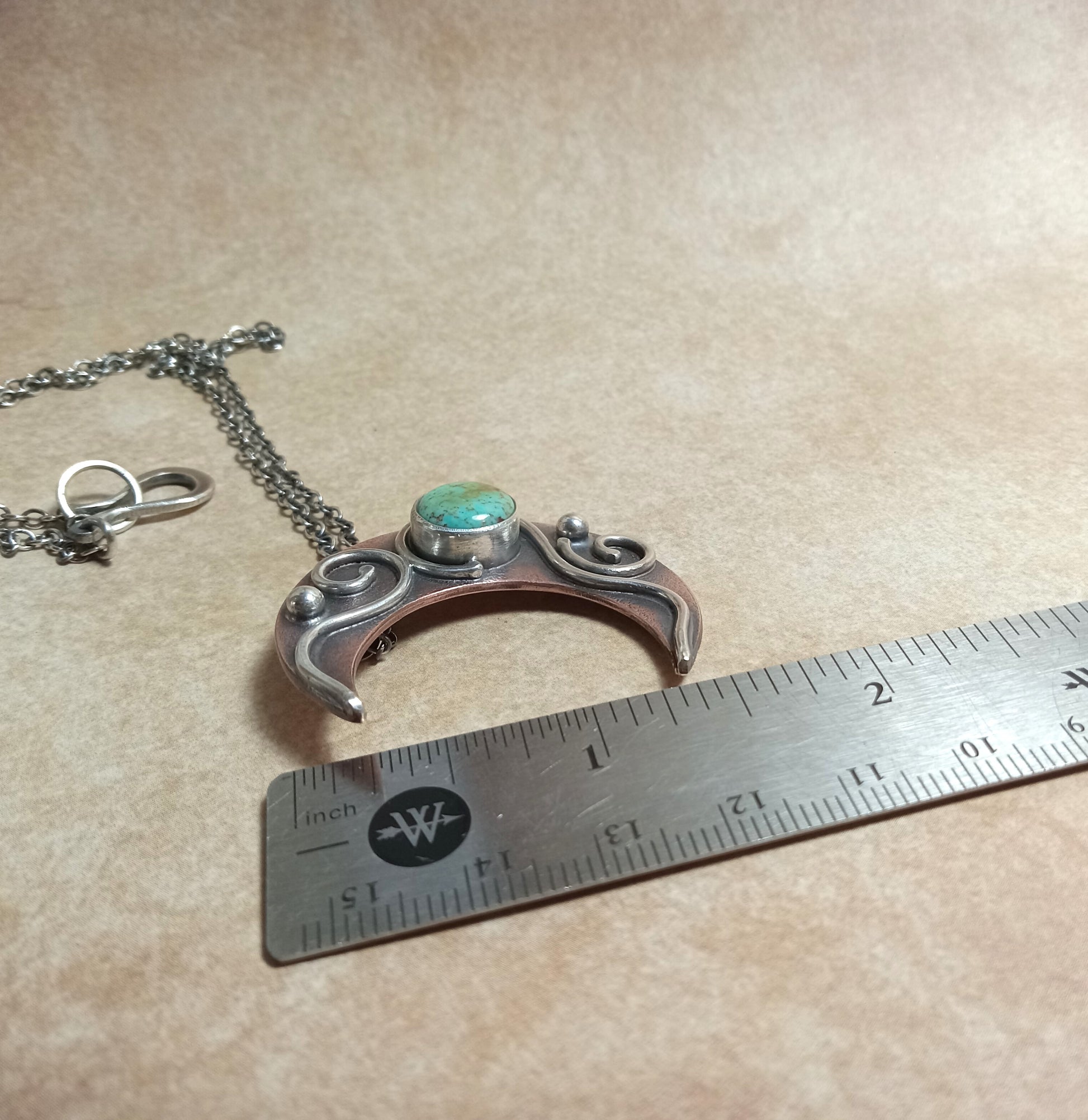Copper and Silver pendant with turquoise stone on a beige surface with a ruler for scale. Rustic jewelry by Mocahete.