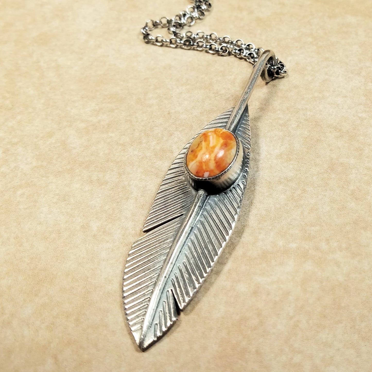Front view of SS feather shaped pendant with an orange spiny oyster on a beige background.