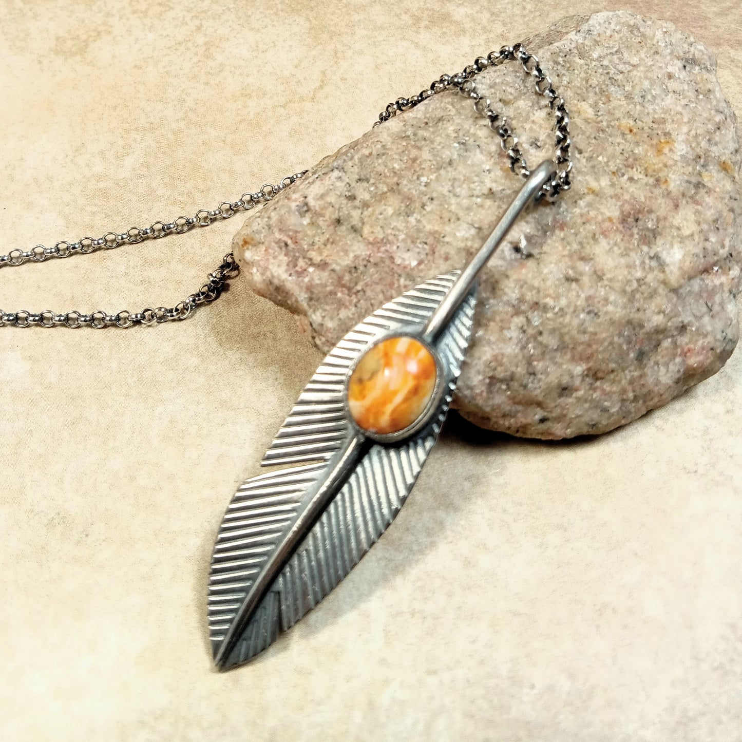 Argentium Silver feather-shaped pendant with an orange spiny oyster cabochon on a SS chain, resting on a rock.