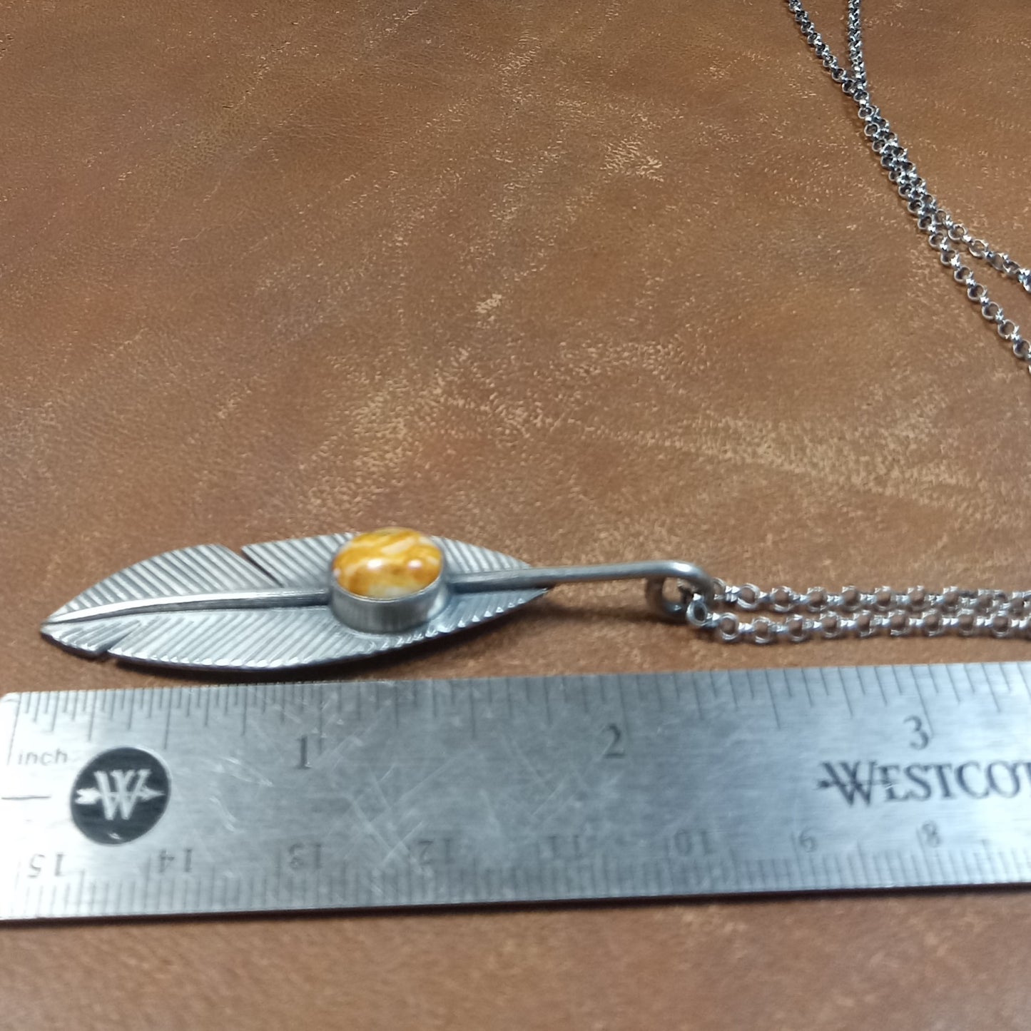 Argentium Silver feather-shaped pendant with orange stone on a brown surface, measured against a ruler