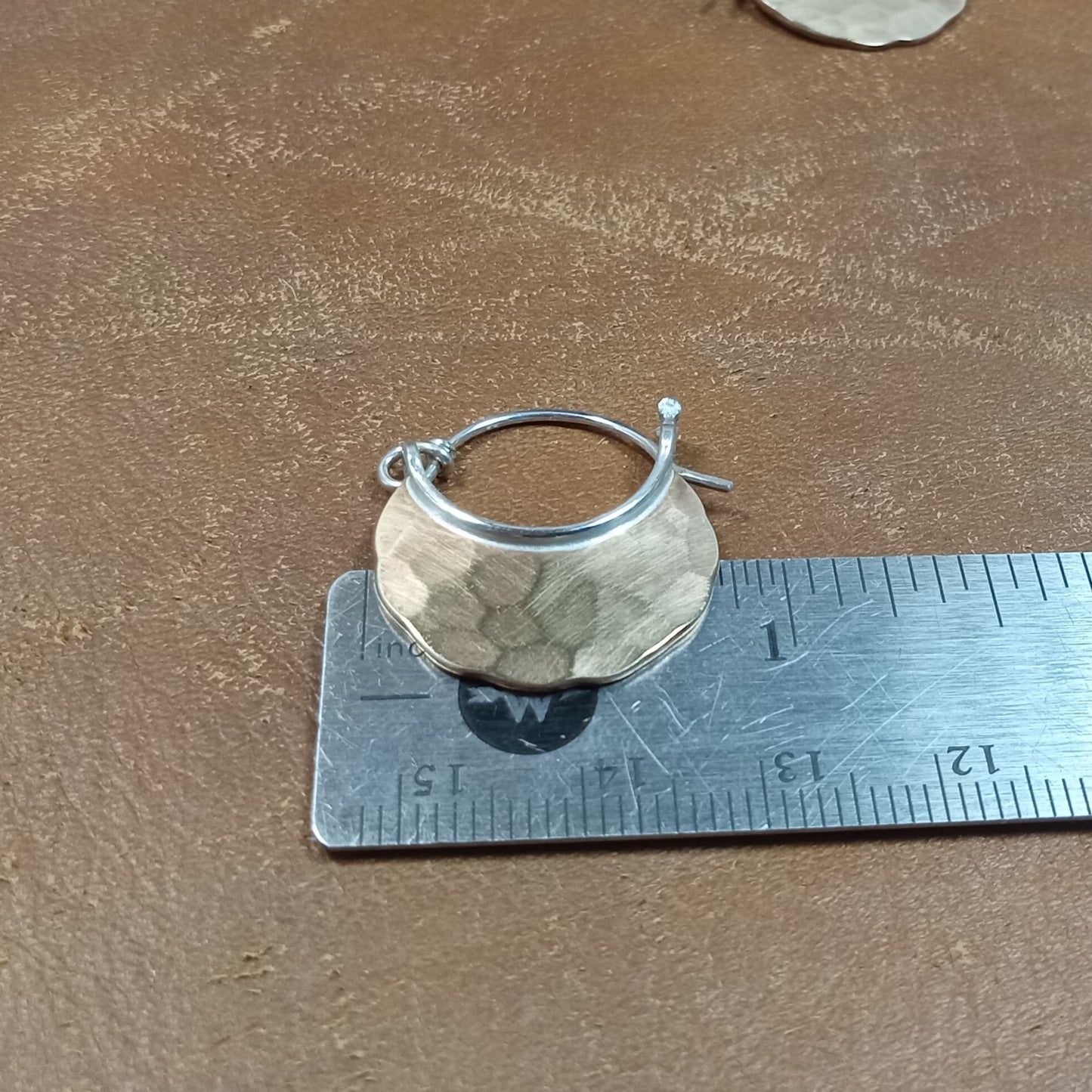 Mixed metal, bronze and silver earring on a ruler for size measurement on a brown surface.