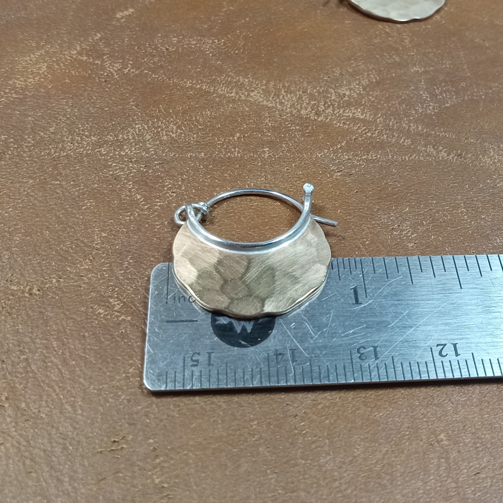 Mixed metal, bronze and silver earring on a ruler for size measurement on a brown surface.