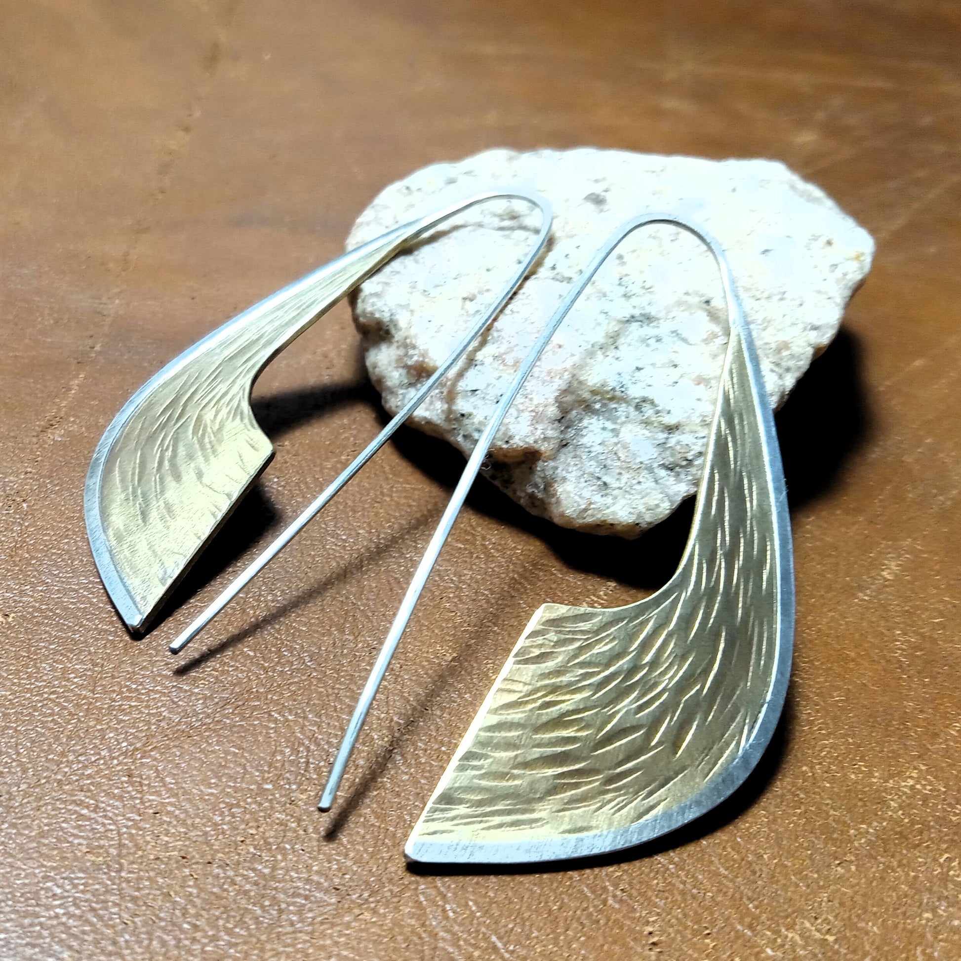 Scimitar shaped Bronze and Silver earrings with textured design on a leather surface with stone.