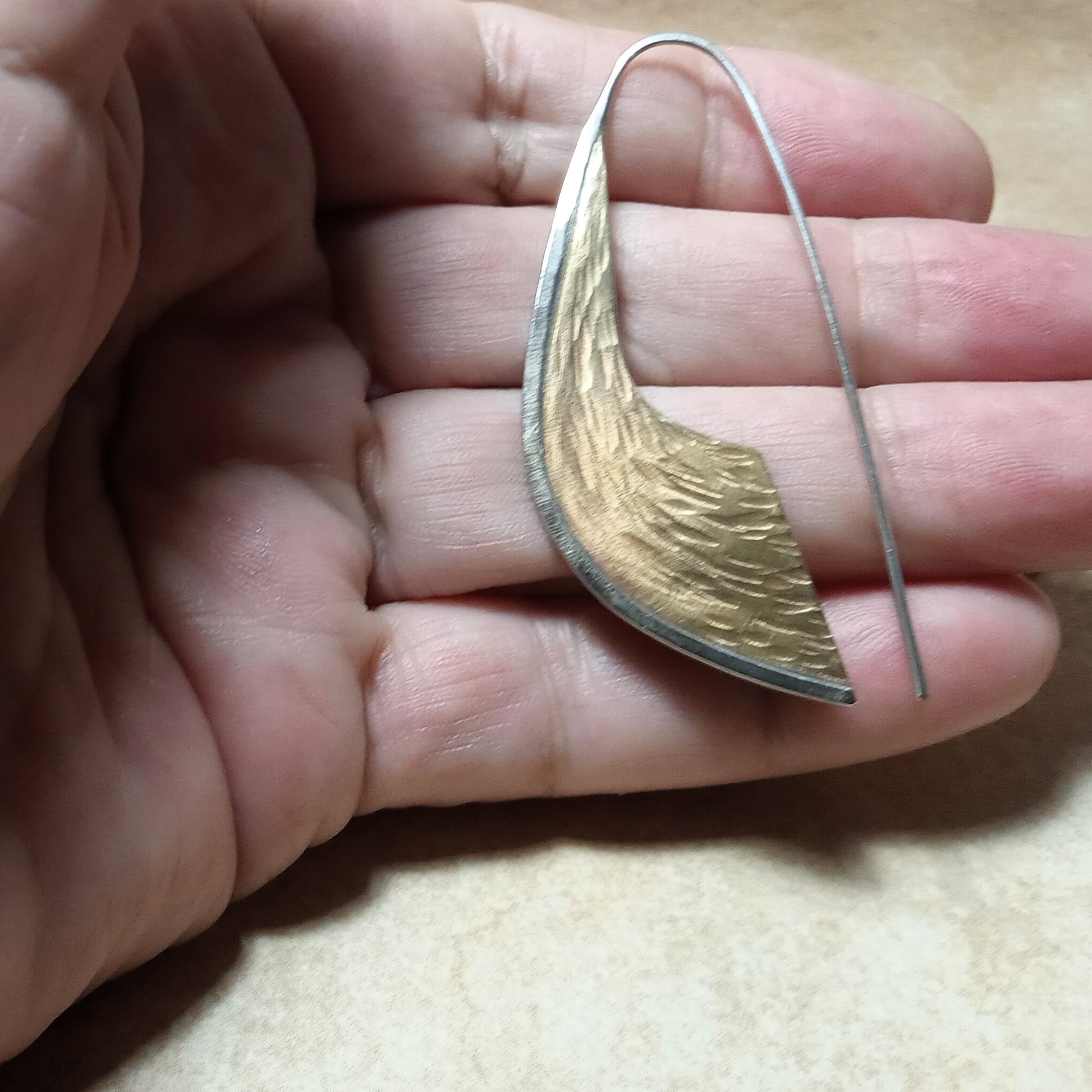 Bronze and silver scimitar shaped earring held on fingers on a neutral background