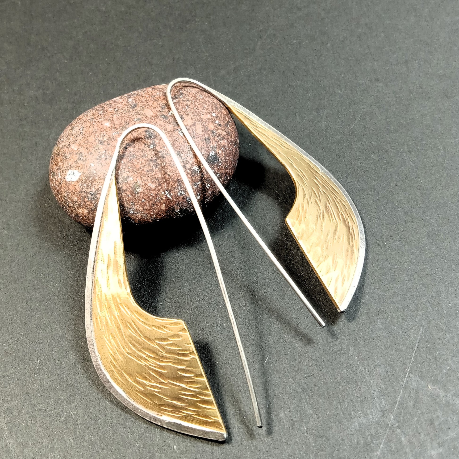 Pair of bold bronze and silver earrings on a dark surface with a stone.