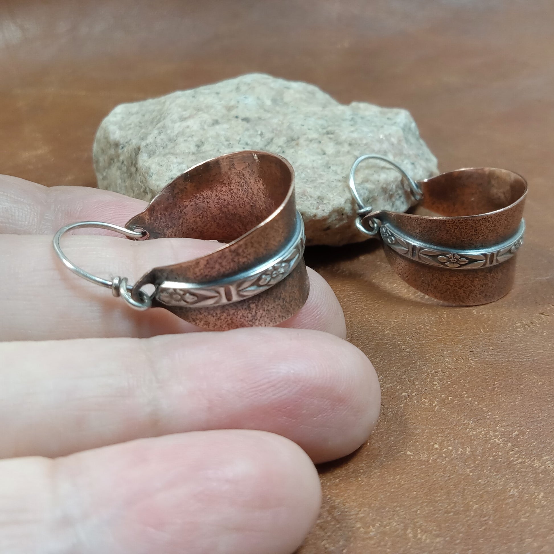 Textured copper hoop earring with sterling silver floral accent band resting on hand with stone and the  other earring in background.