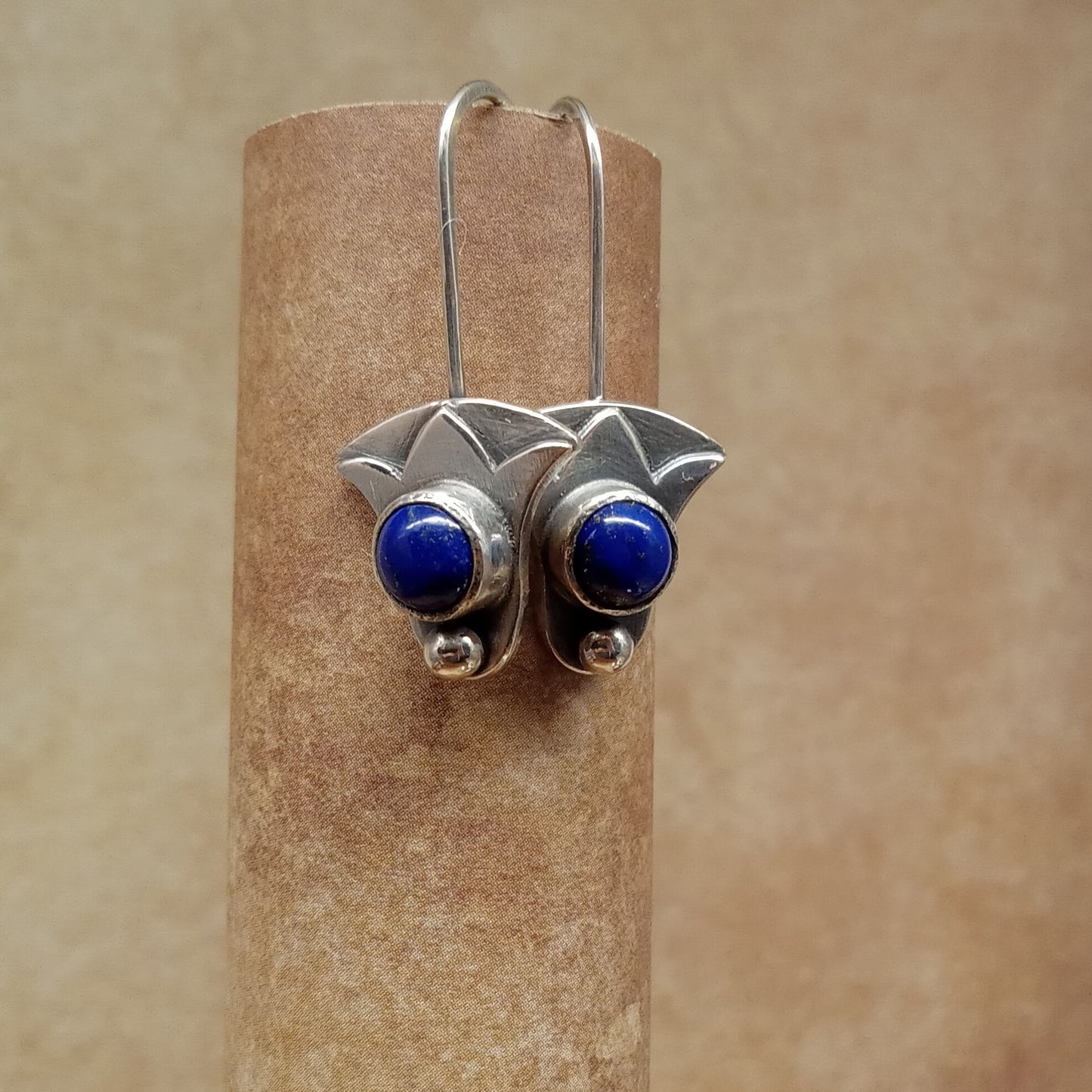 Silver earrings with blue  lapis stones on a cylindrical brown stand against a beige background