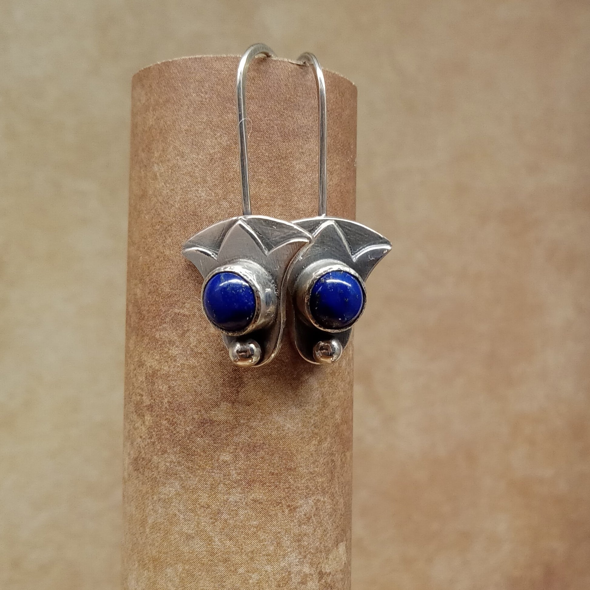 Silver earrings with blue  lapis stones on a cylindrical brown stand against a beige background