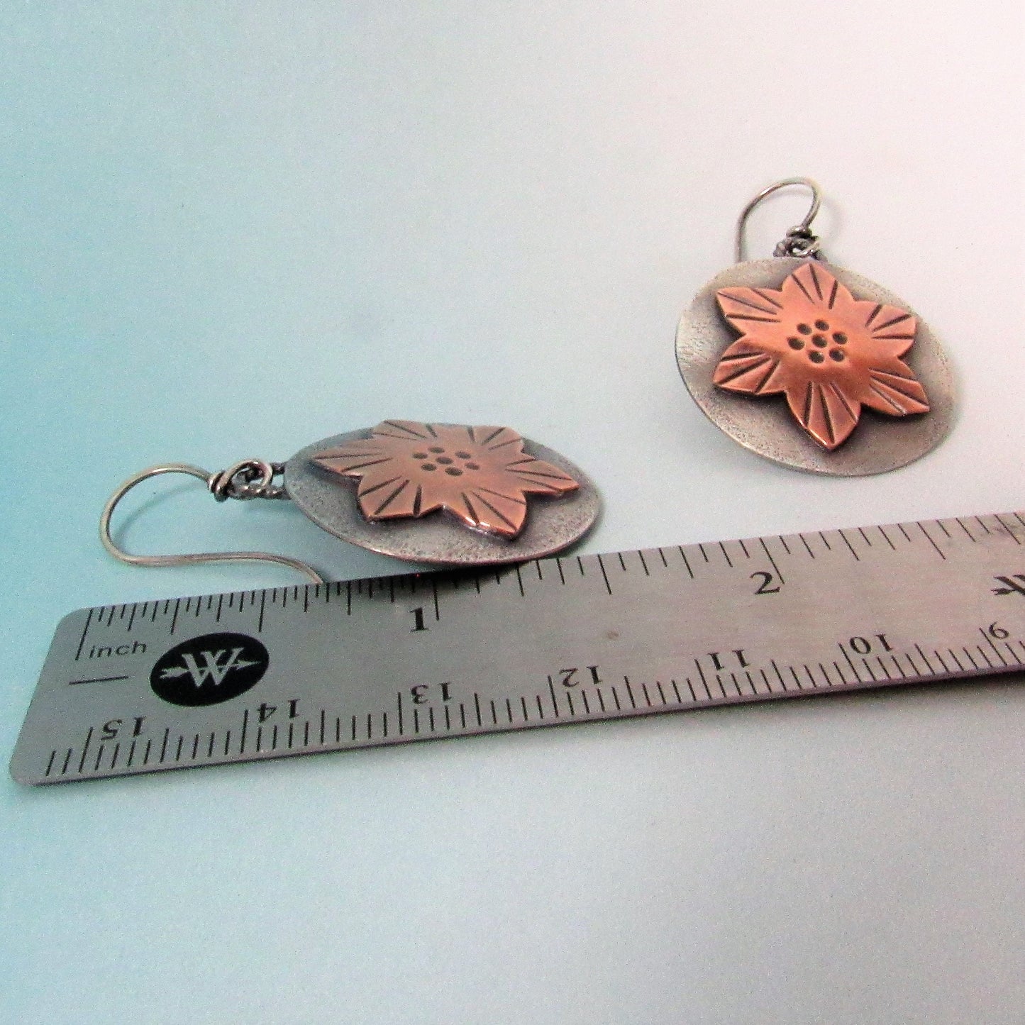 Copper flower-shaped earrings on a ruler for scale against a light blue background