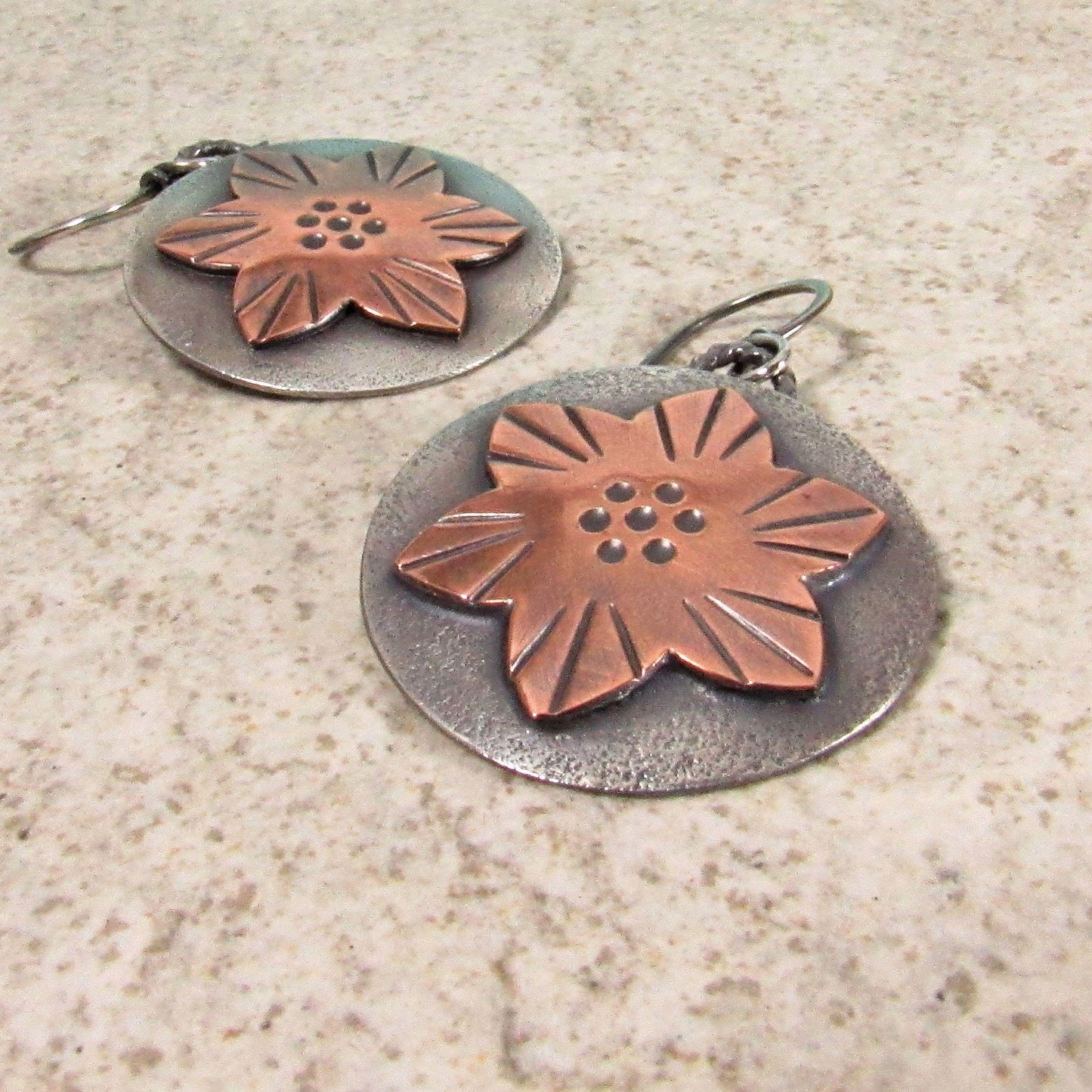 Artisan dangle earrings with warm copper and cool silver contrast in a star-shaped flower design. On textured beige surface.