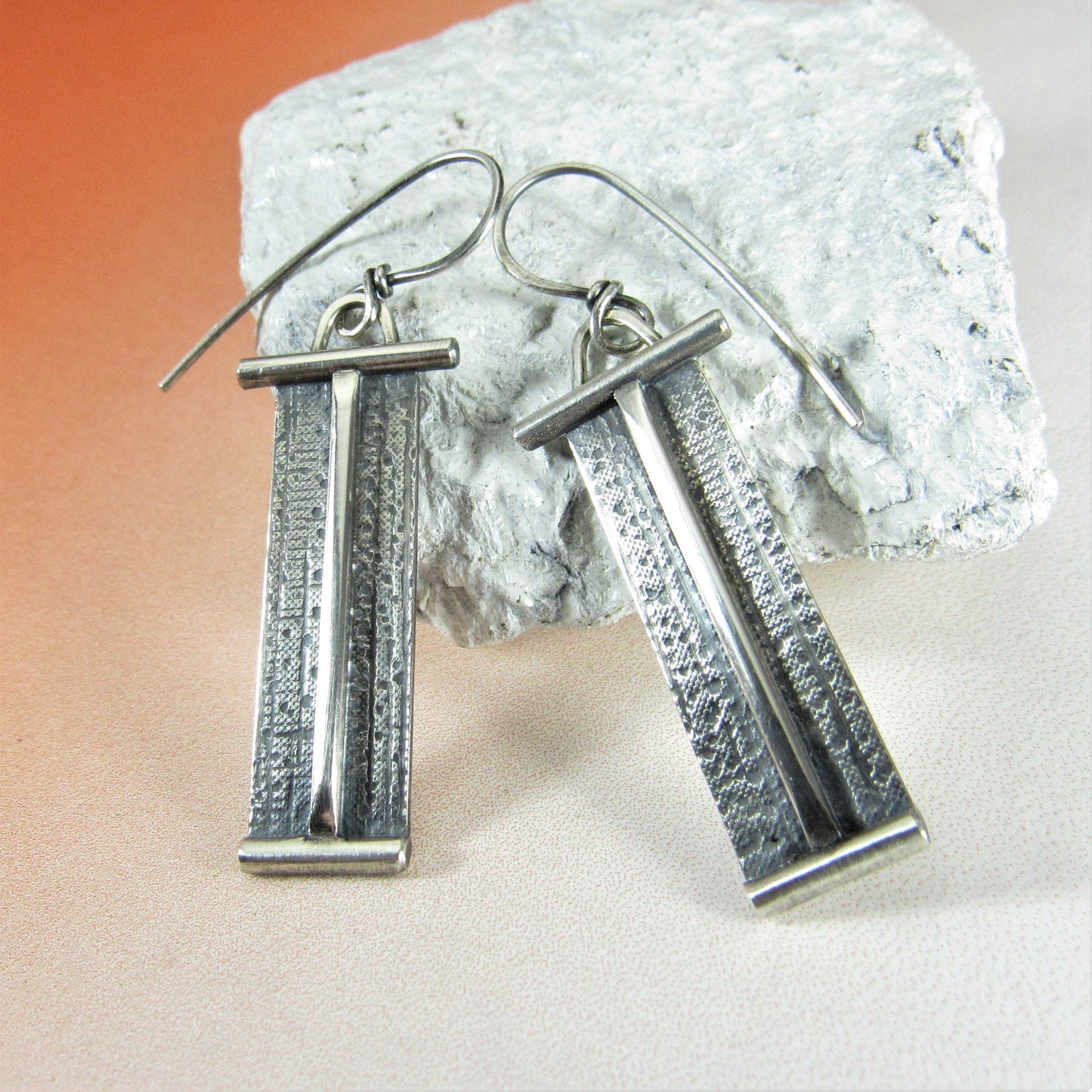 Silver Zen earrings with textured bars against white stone and orange paper.