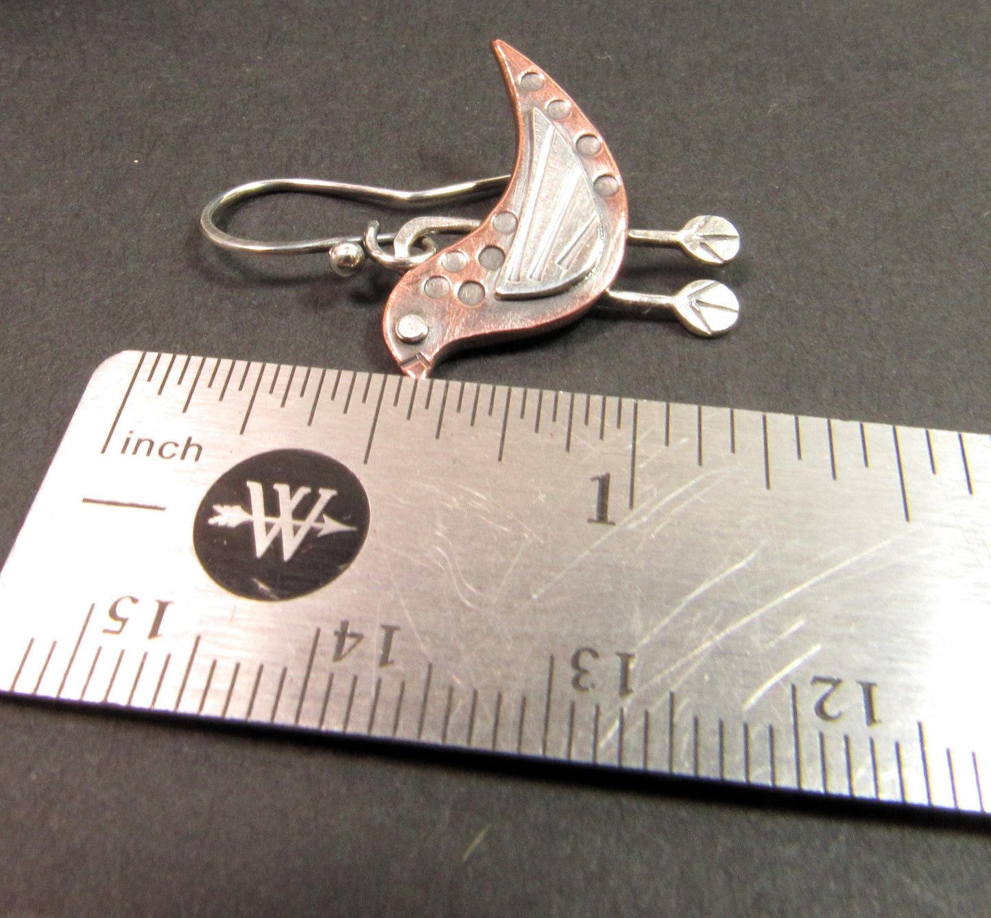 Playful copper and silver bird-shaped earrings with hand-stamped details and polished patina. Earring next to ruler for scale.
