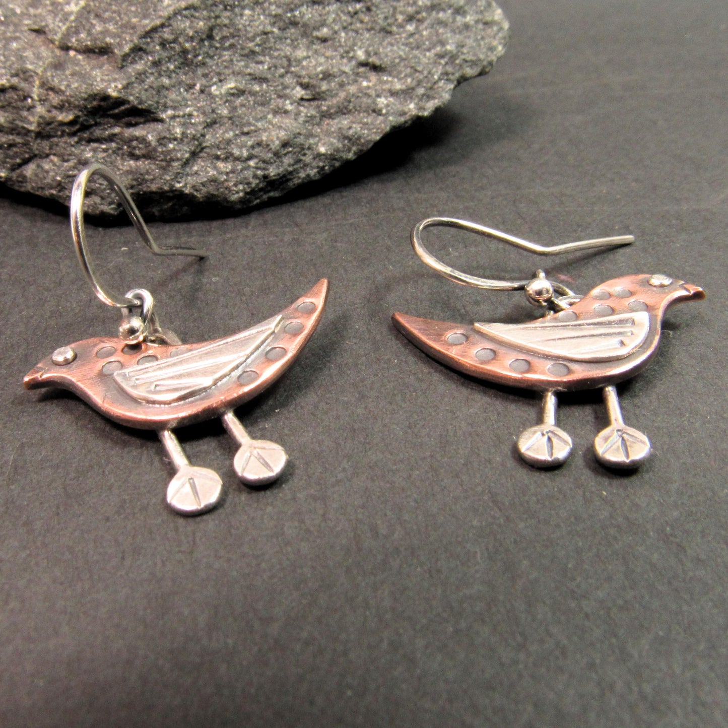 Mixed metal bird earrings featuring oxidized copper bodies and silver eyes, wings, and stamped feet. On dark background with rock.