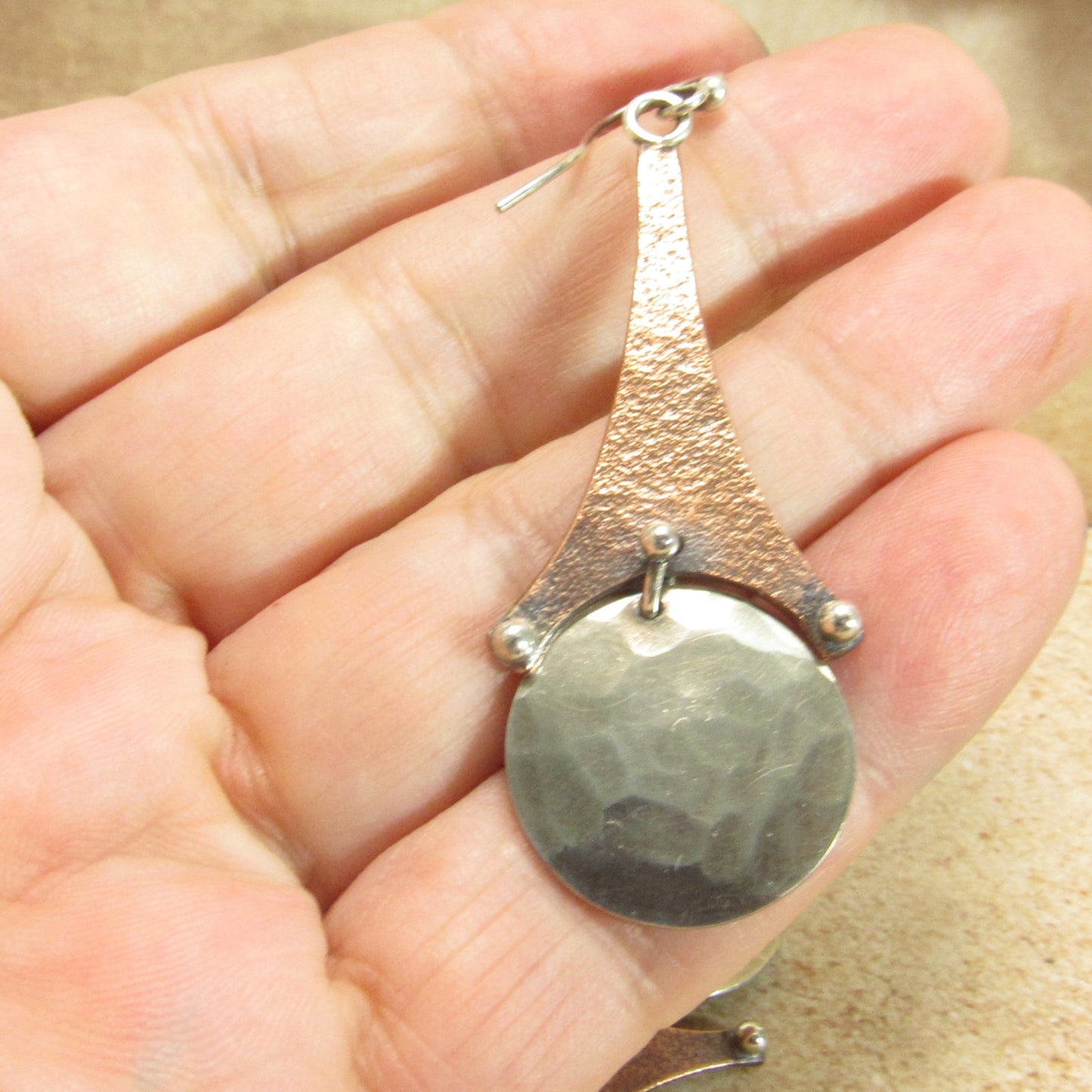Argentium Silver and copper earring with textured design held between fingers on a beige background.
