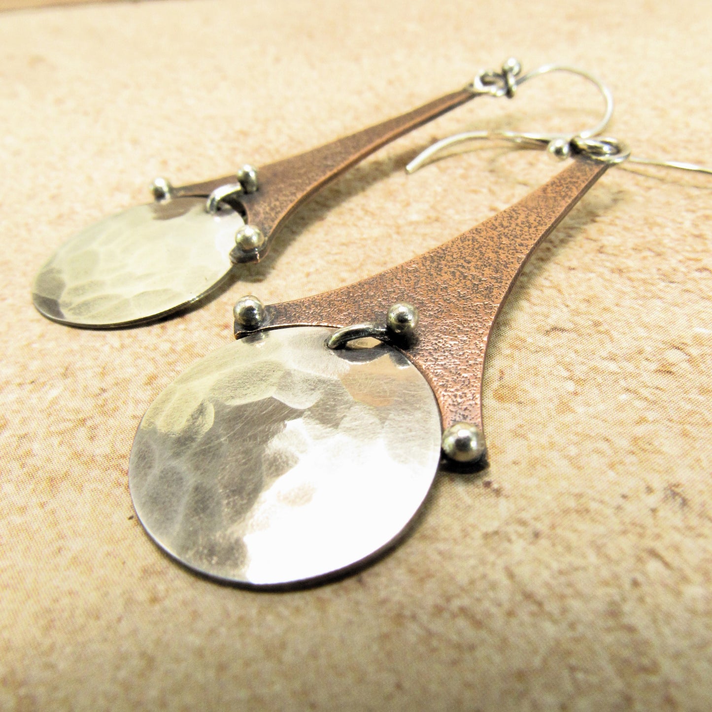 Handcrafted mixed metal earrings featuring oxidized copper bars suspending hammered Argentium silver disks with silver ball accents, artisan made by Mocahete. Photo on textured beige background.