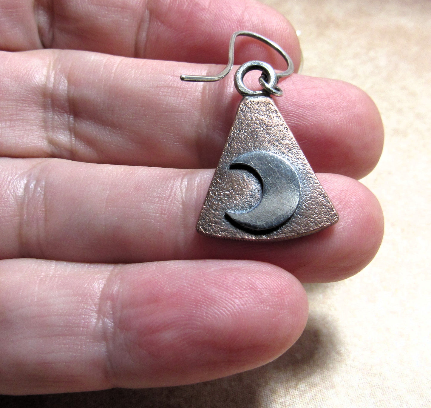 Triangular earrings in silver and copper with a moon design held on hand.