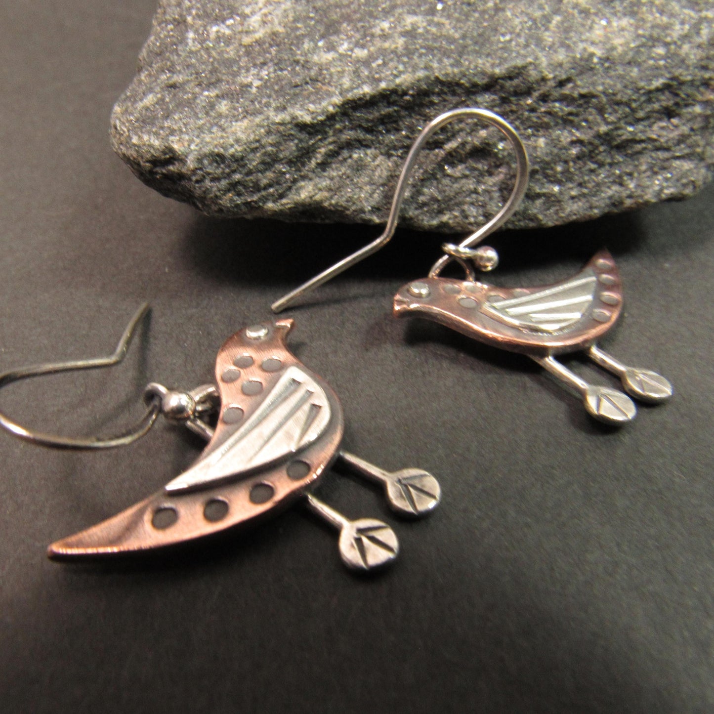 Copper and silver bird-shaped earrings on a dark stone surface.