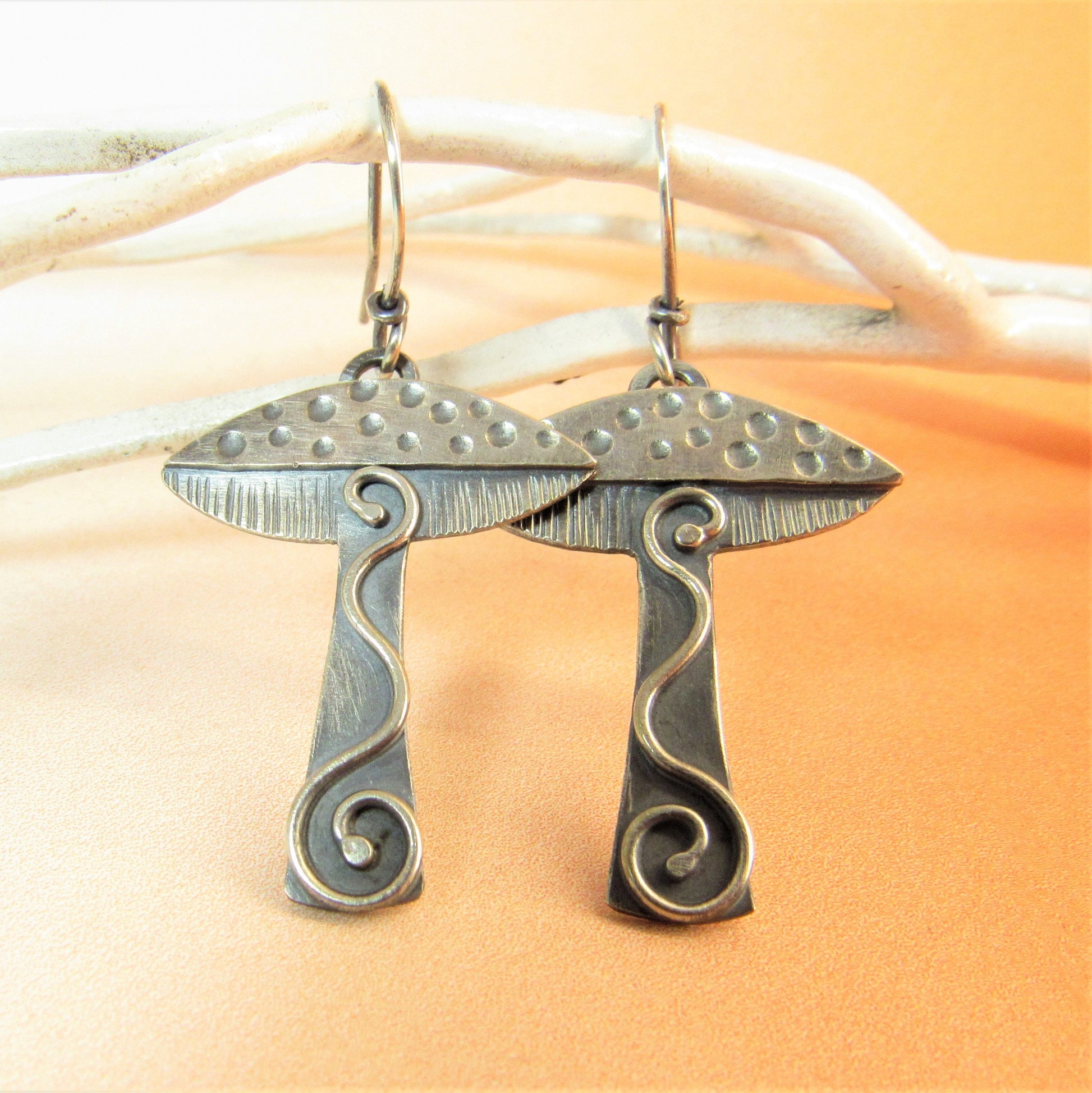 Handcrafted silver mushroom earrings on whte twig and orange background.