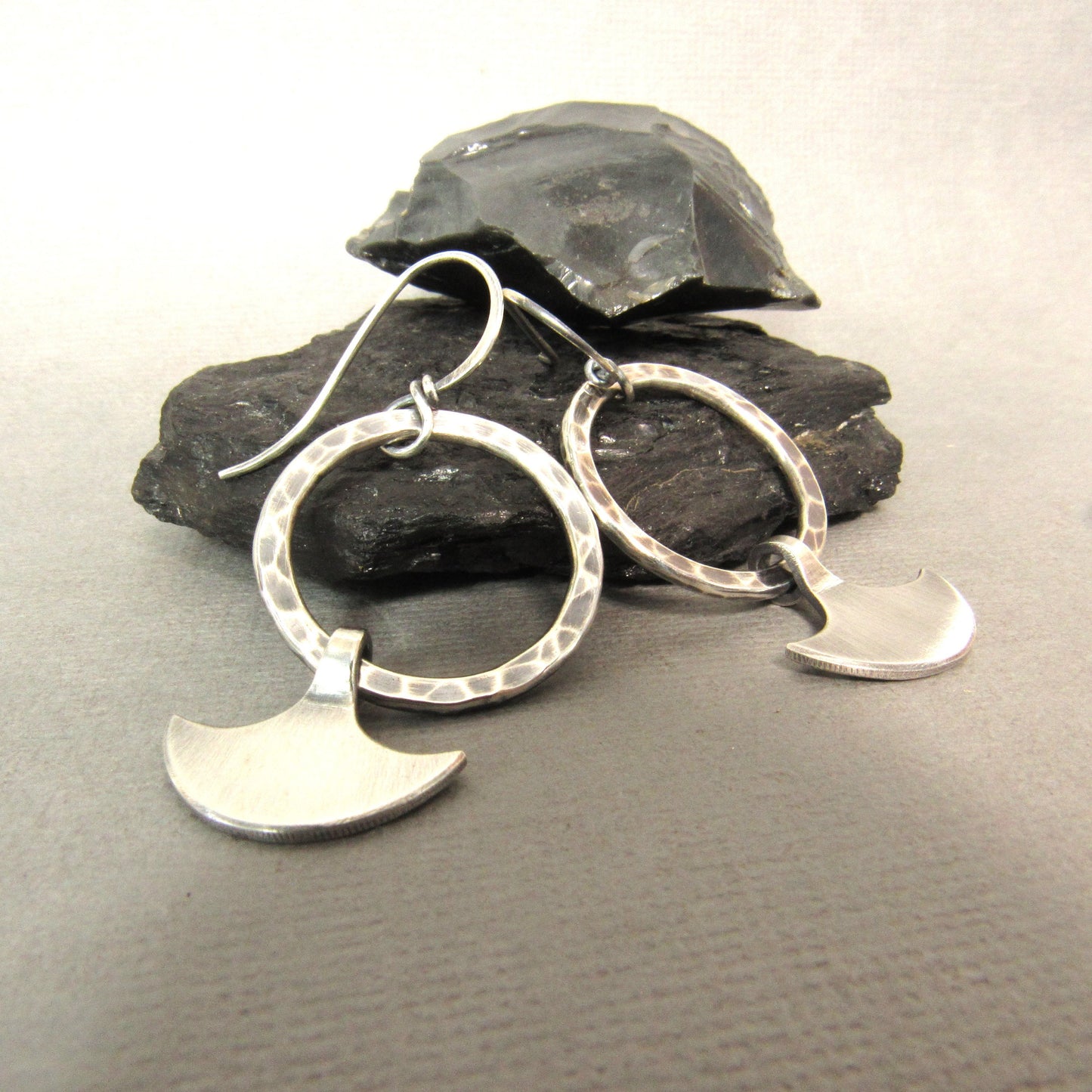 Silver Hoop dangle earrings with ax charm against rocks on grey background.