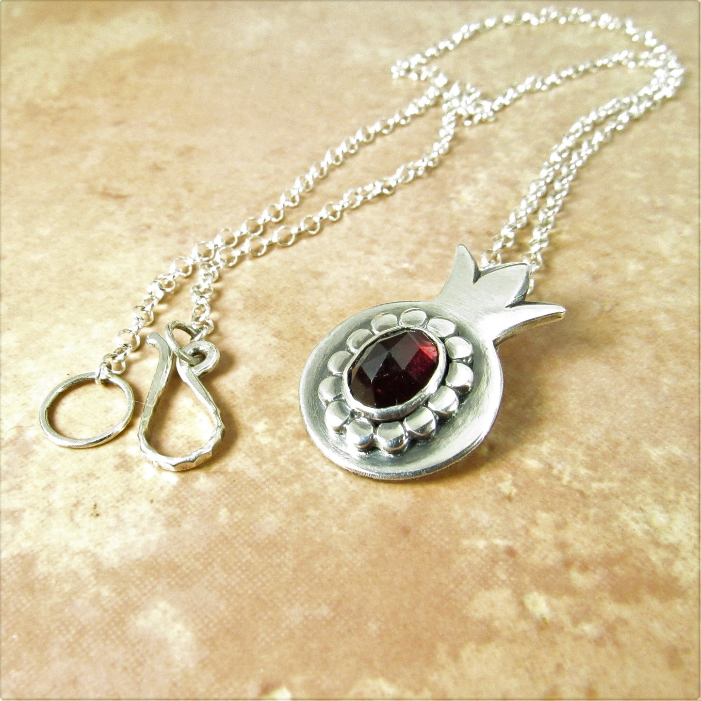 Silver  pomegranate pendant necklace  a garnet gemstone, shows clasp and chain  on a beige background