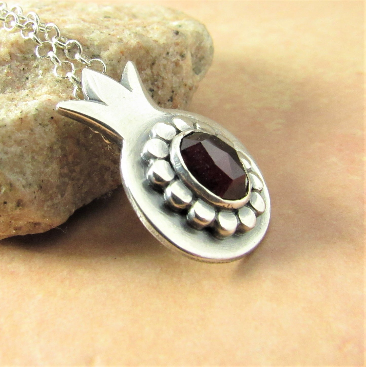 Side view pomegranate pendant, argentium sterling silver and faceted garnet photo on stone and beige background.