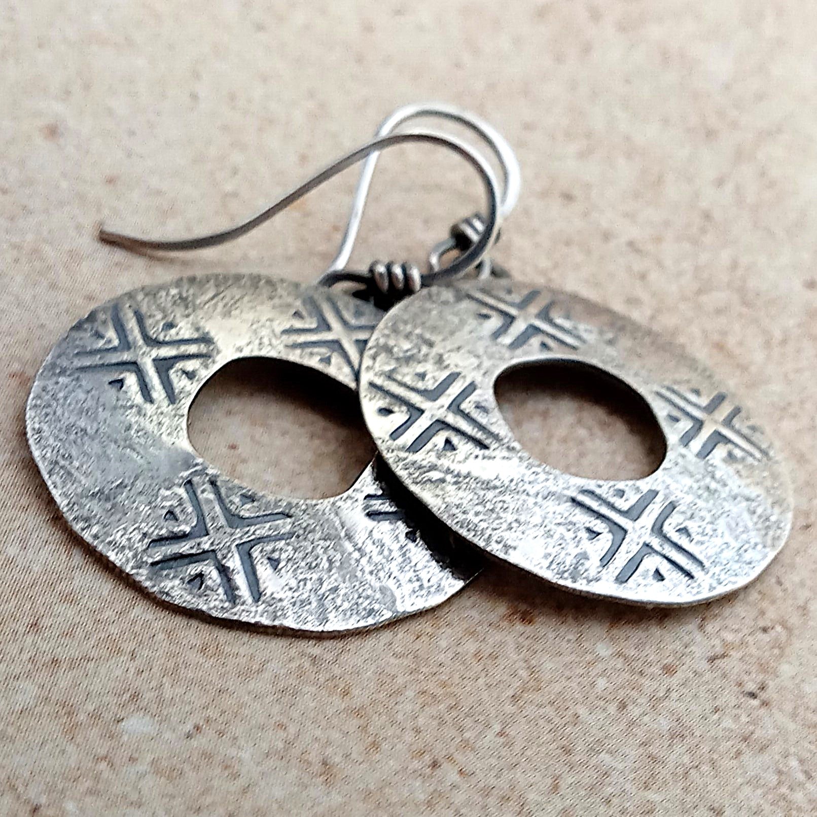 Handcrafted argentium SS coin style pair of silver earrings with geometric patterns on a beige background.
