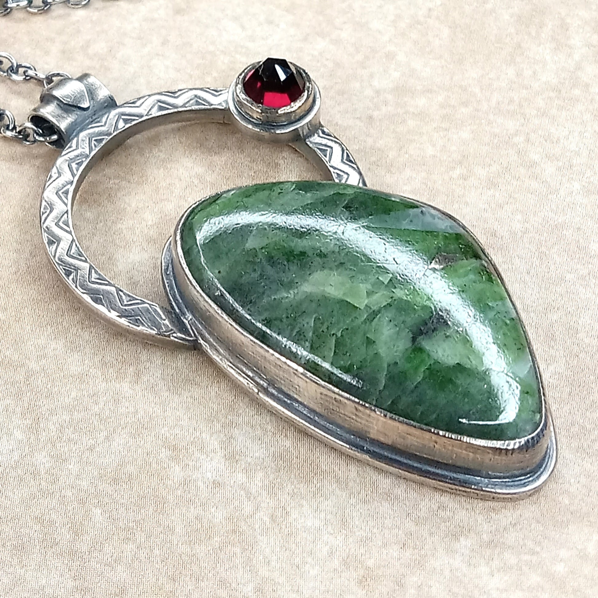 Green stone pendant with silver and red gemstone on a beige fabric background