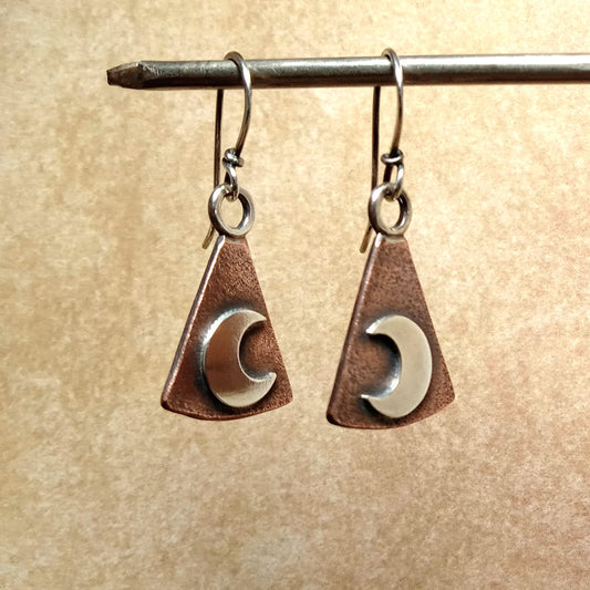 Pair of earrings with triangular copper and silver crescent moon design on a beige background.