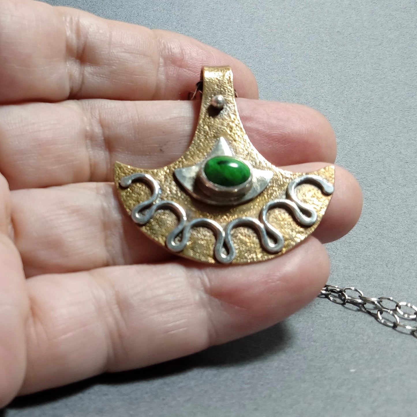 Bronze and silver pendant ax style with a green maw sit sit gemstone held in a hand on a gray background