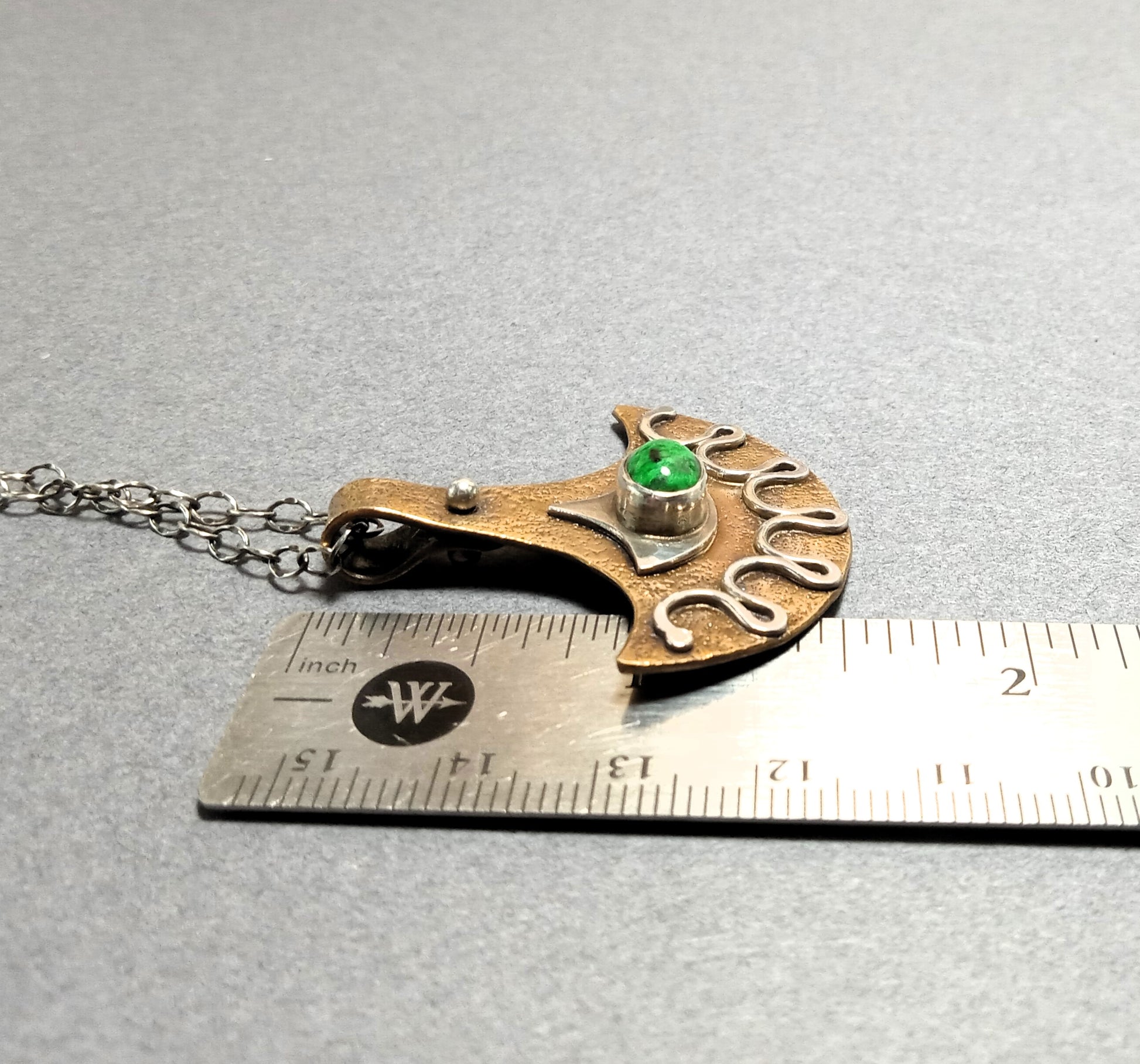 Decorative tribal style pendant with a green maw sit sit gemstone on a chain, measured against a ruler.